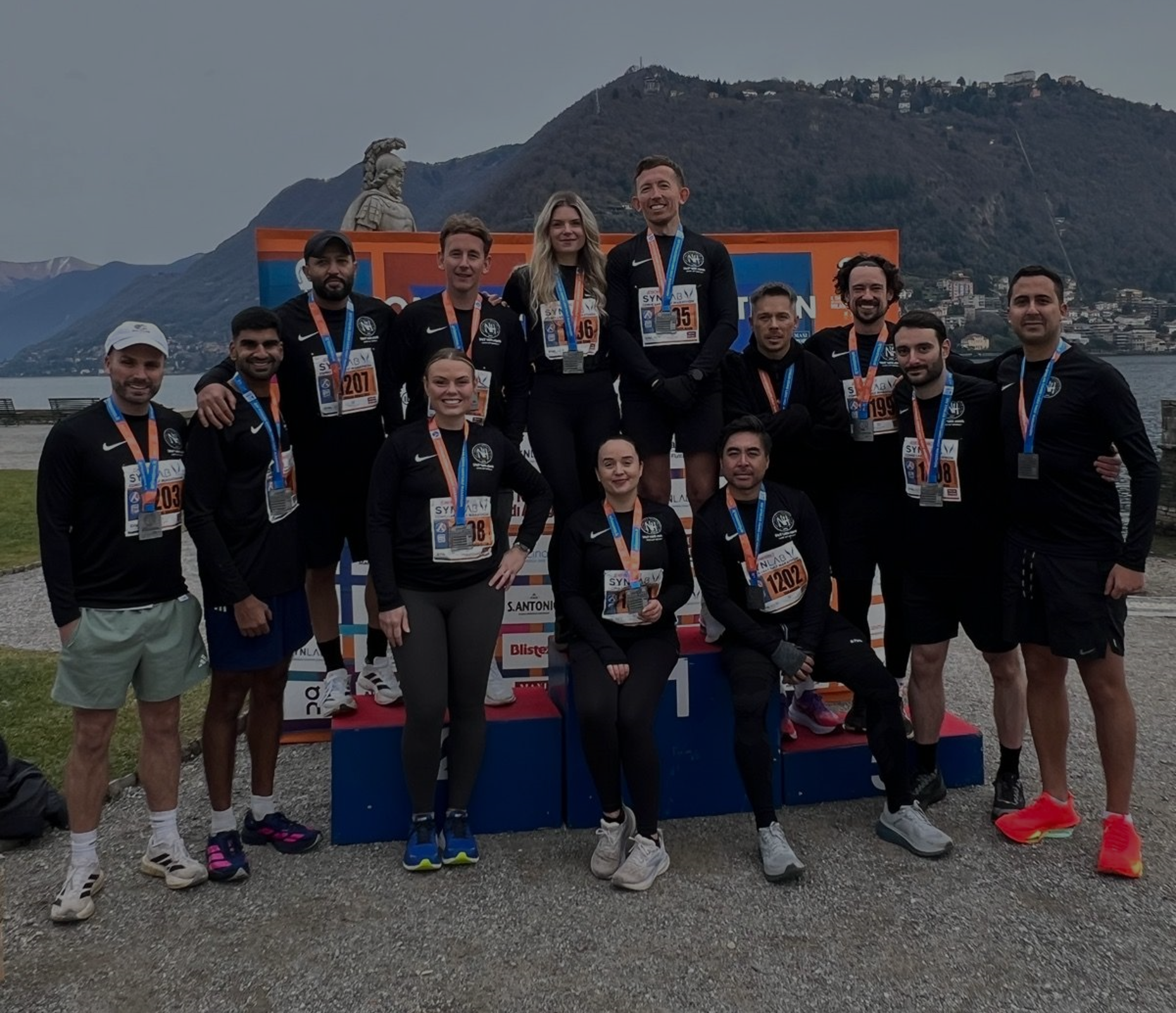 Group of runners in athletic gear posing for a photo at a race event near a lake with mountains in the background, some standing on a tiered podium, wearing medals and race bibs.