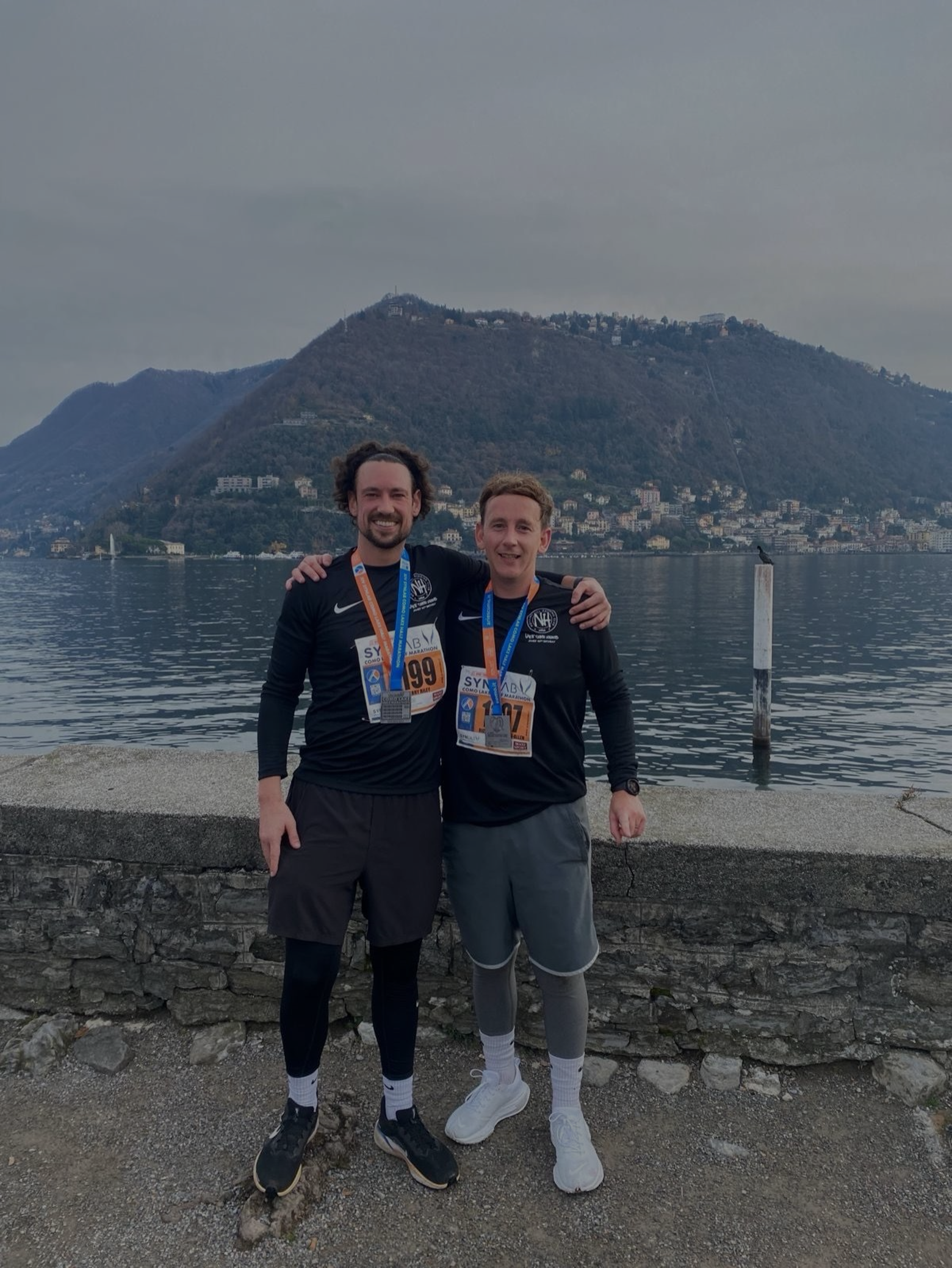 Two men standing together by a body of water, wearing running clothes and medals, with a mountain and town in the background.