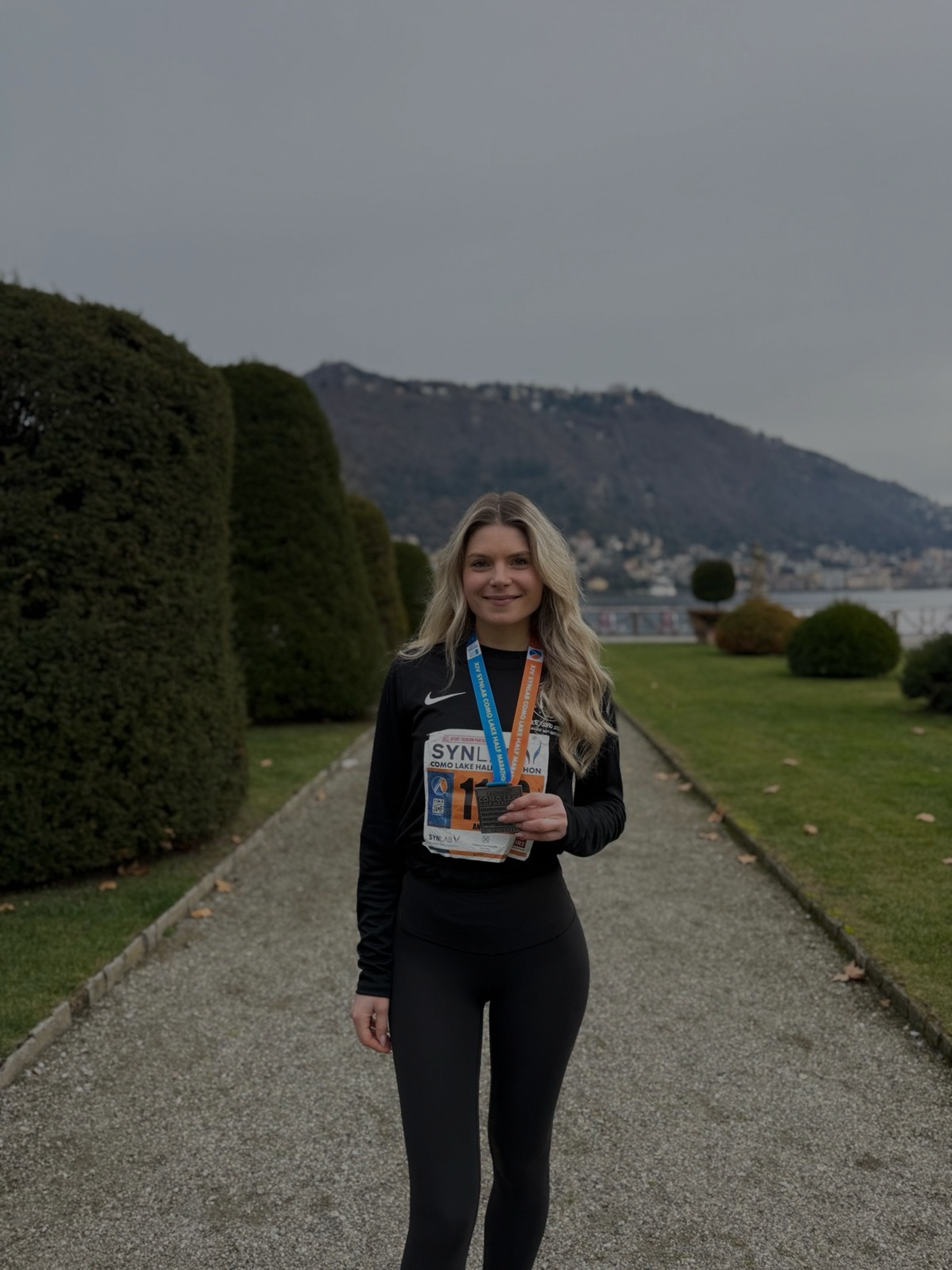 A young woman in athletic clothing holding a race bib and medals, standing on a gravel path with trimmed bushes and a body of water in the background.
