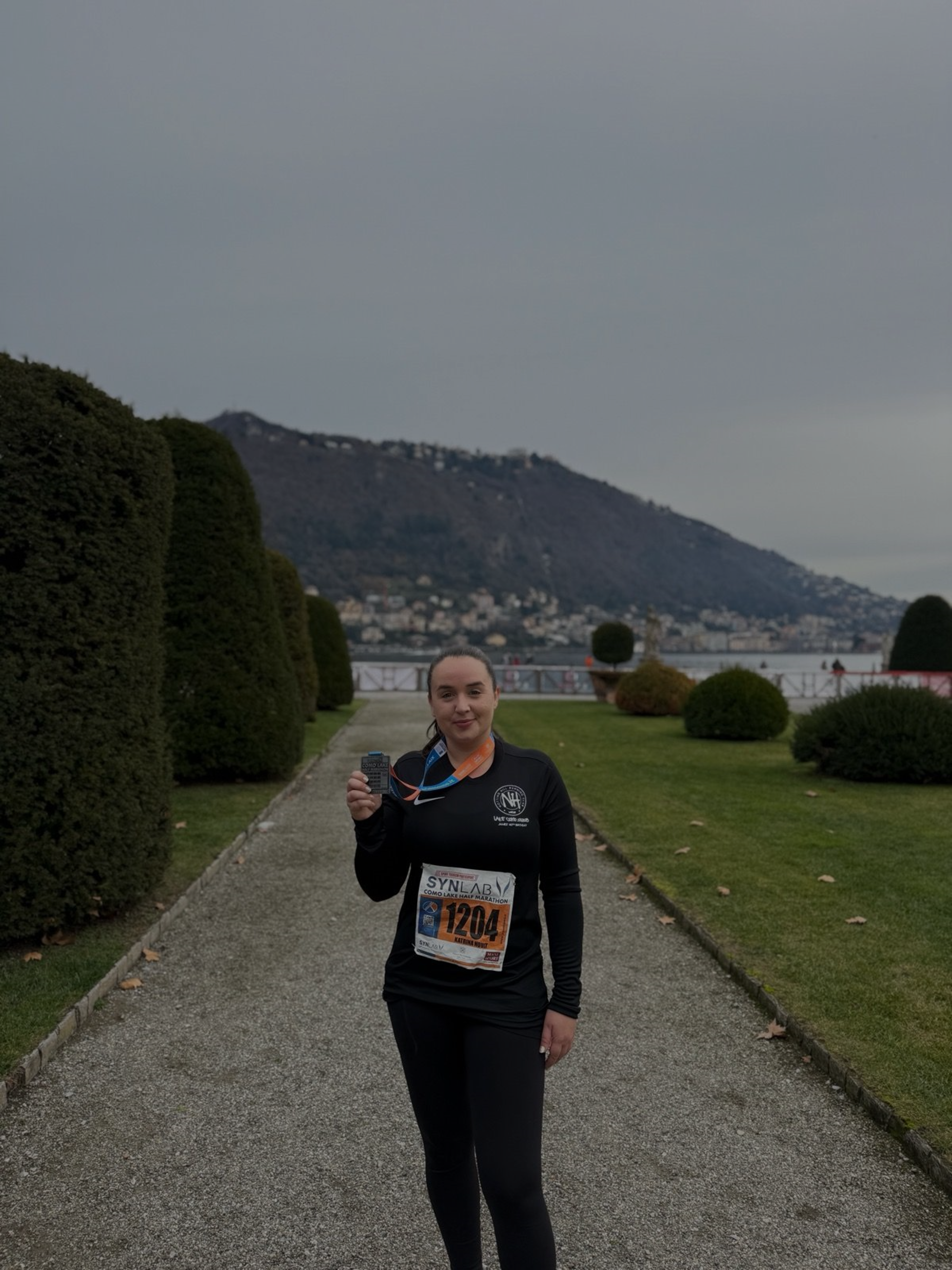 A woman standing on a gravel path holding a race medal, wearing a black athletic outfit with a bib number 1204, in a garden with trimmed hedges, overlooking a body of water and hills in the background.