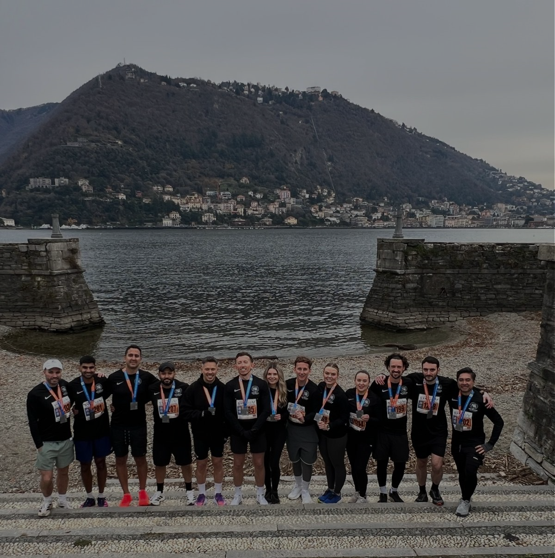 Group of runners with medals posing on a waterfront promenade near a lake with mountains in the background.