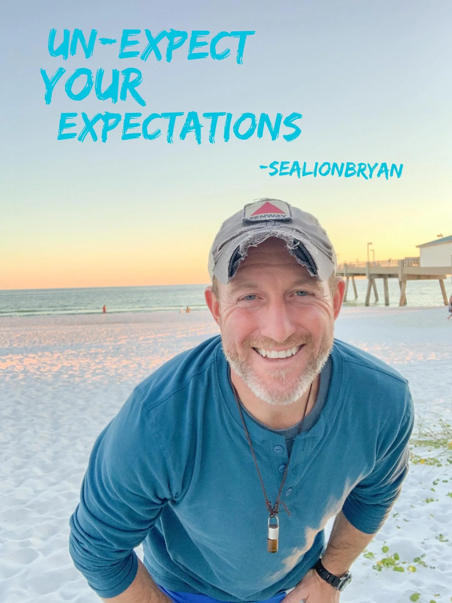 Smiling man wearing a baseball cap and blue shirt on a beach at sunset
