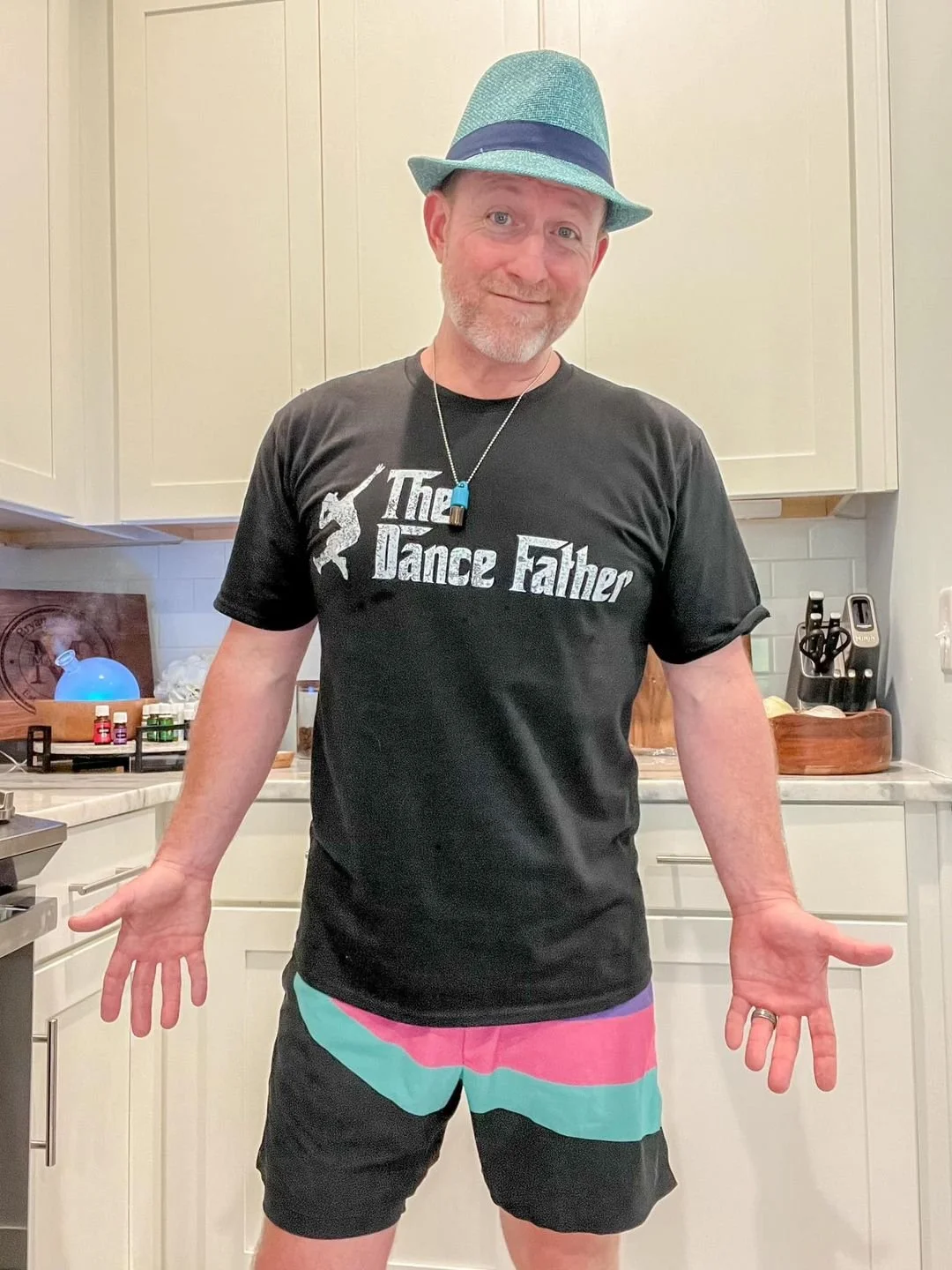 A man wearing a teal fedora hat, a black T-shirt with the words 'The Dance Father' and a dancer silhouette, and colorful shorts. He is standing in a kitchen with white cabinets, surrounded by kitchen utensils and decor.