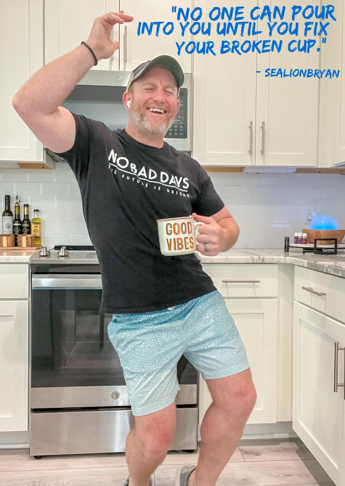 A man standing in a kitchen, smiling and posing with one arm raised. He is holding a mug that says 'Good Vibes' and wearing a black t-shirt with the words that seem to read 'NOBADDAYS THE FUTURE IS BRIGHT,' light blue shorts, and a baseball cap.