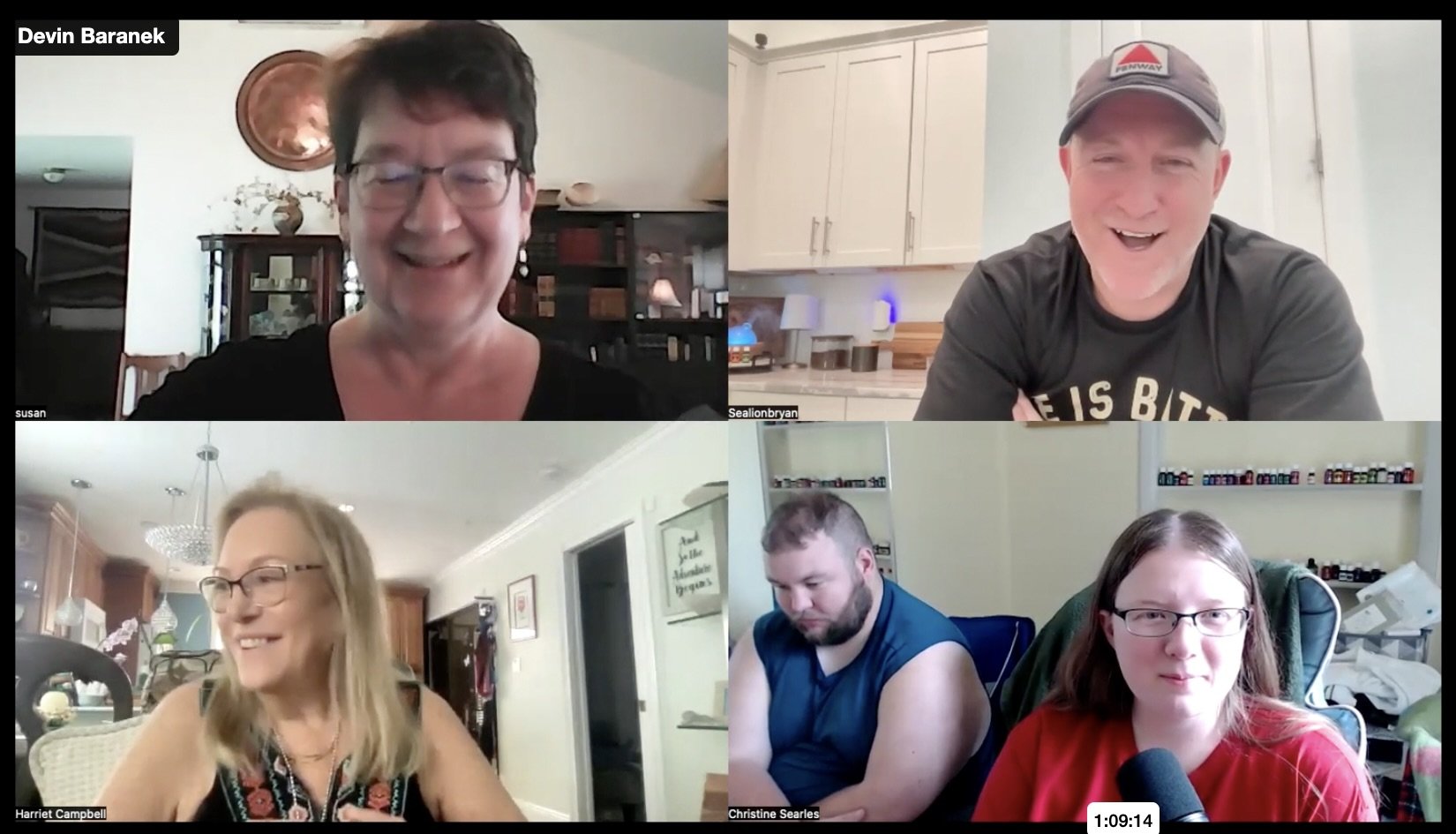A screenshot of a video call with five participants, each in separate environments. The top left is a woman with short dark hair and glasses, smiling in a room with a cabinet and a copper tray on the wall. The top right is a man with a cap, smiling and wearing a black T-shirt, in a kitchen. The bottom left is a woman with glasses and long blonde hair, smiling in a living room with hanging lights. The bottom middle is a man with a beard, looking down, in a room with shelves and framed pictures. The bottom right is a woman with glasses and long brown hair, sitting in a room with shelves and a microphone, looking directly at the camera.