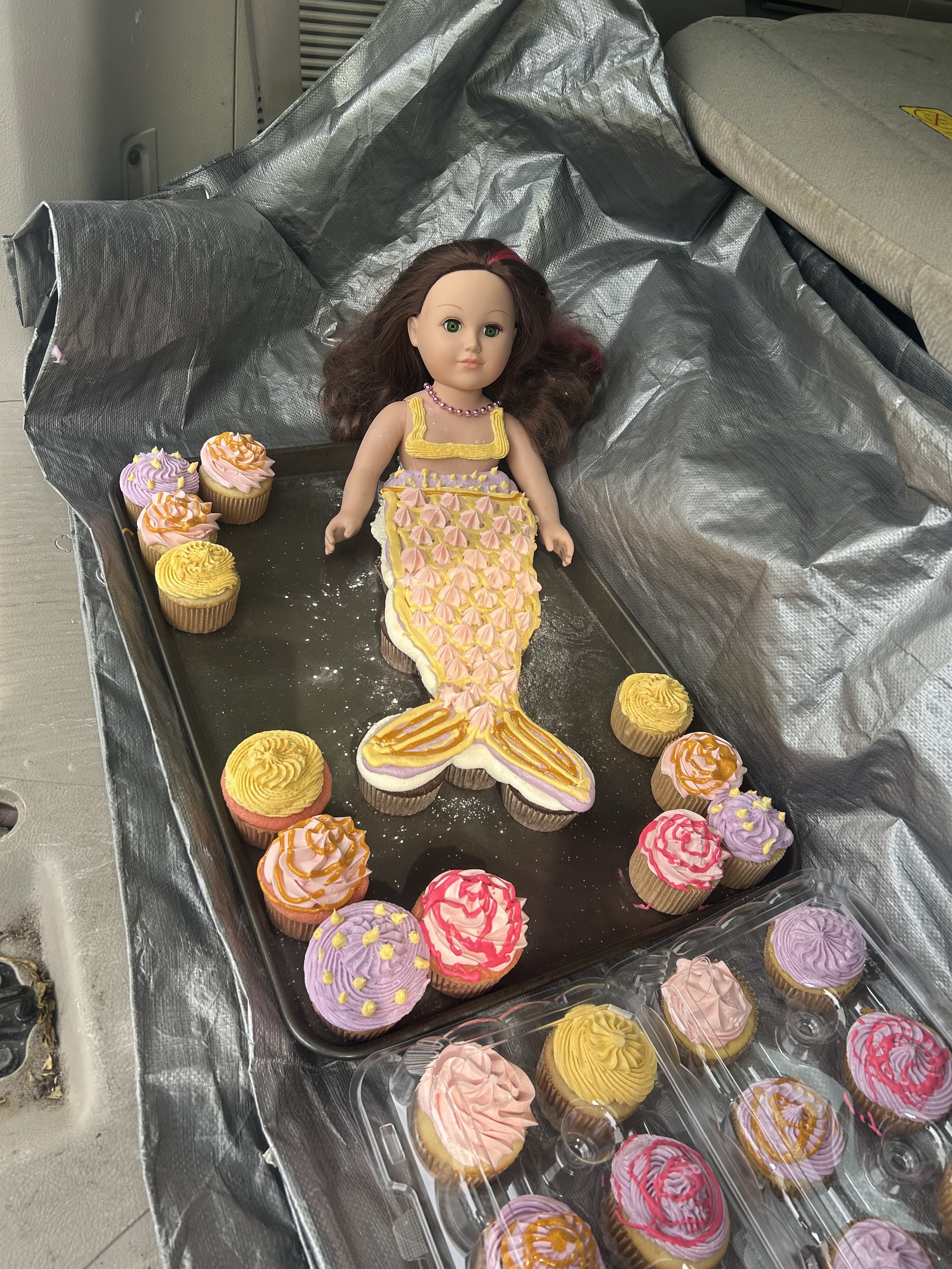 A doll with long brown hair and a yellow mermaid tail decorated with pink and yellow icing is lying on a baking tray surrounded by colorful cupcakes with pink, yellow, and purple icing.