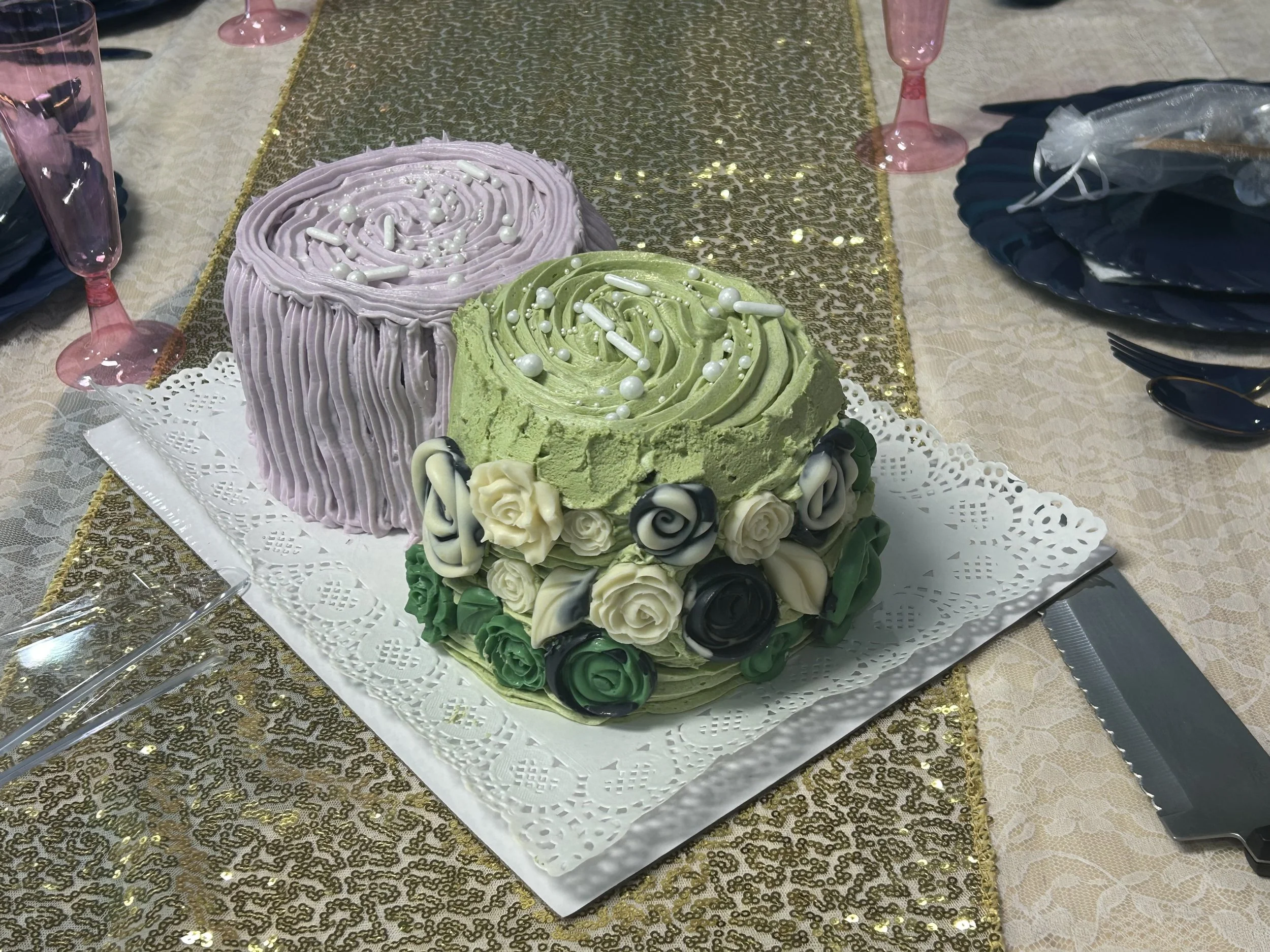 A layered cake with purple and green frosting decorated with white, black, and green roses and pearls, on a decorative paper placed on a gold and cream tablecloth at a celebration.