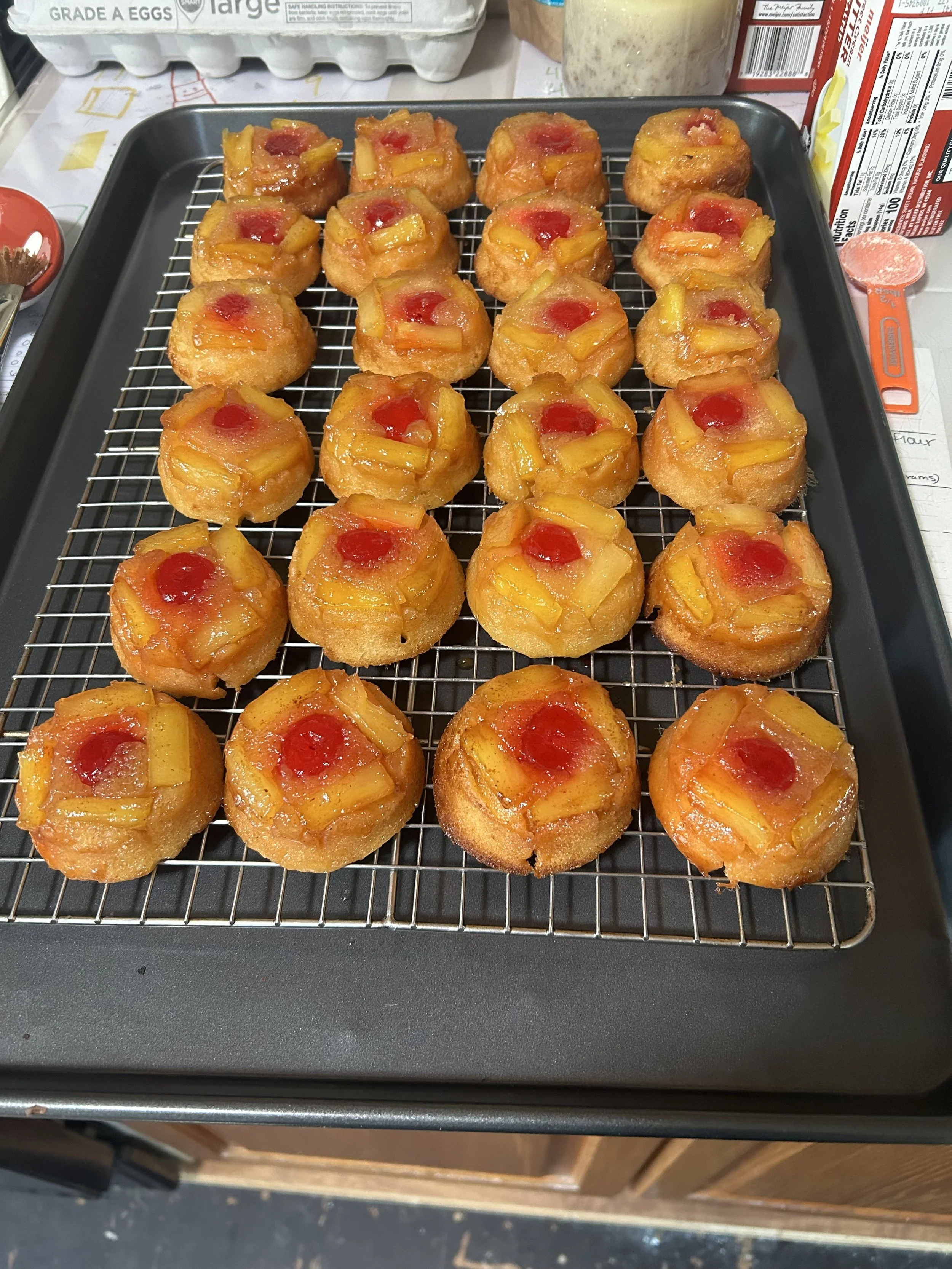 A tray of freshly baked pineapple upside-down cupcakes with a cherry on top, arranged on a wire rack.