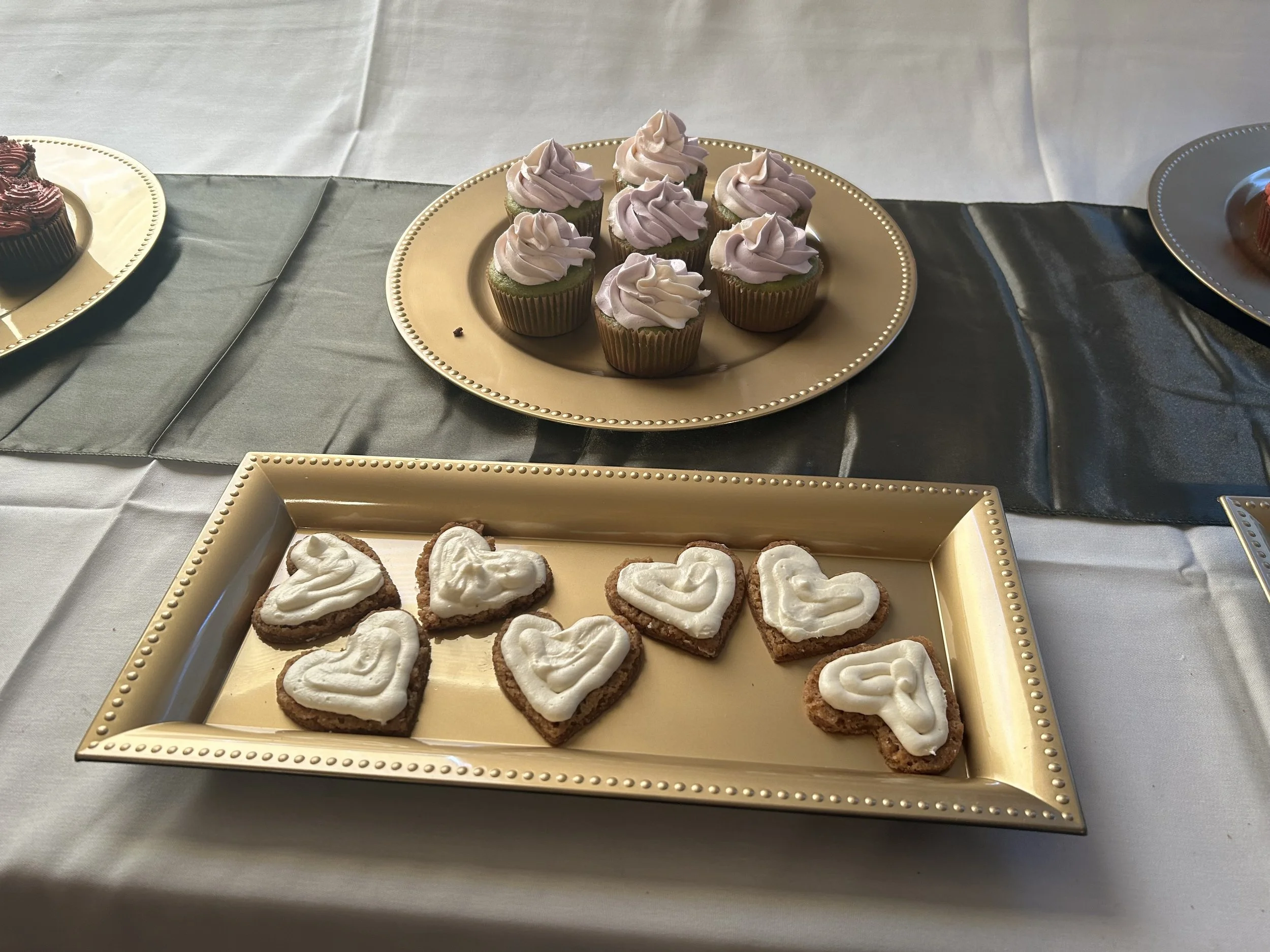 Plate of heart-shaped cookies with white icing and a plate of cupcakes with pink frosting on a table with a gray and white tablecloth.
