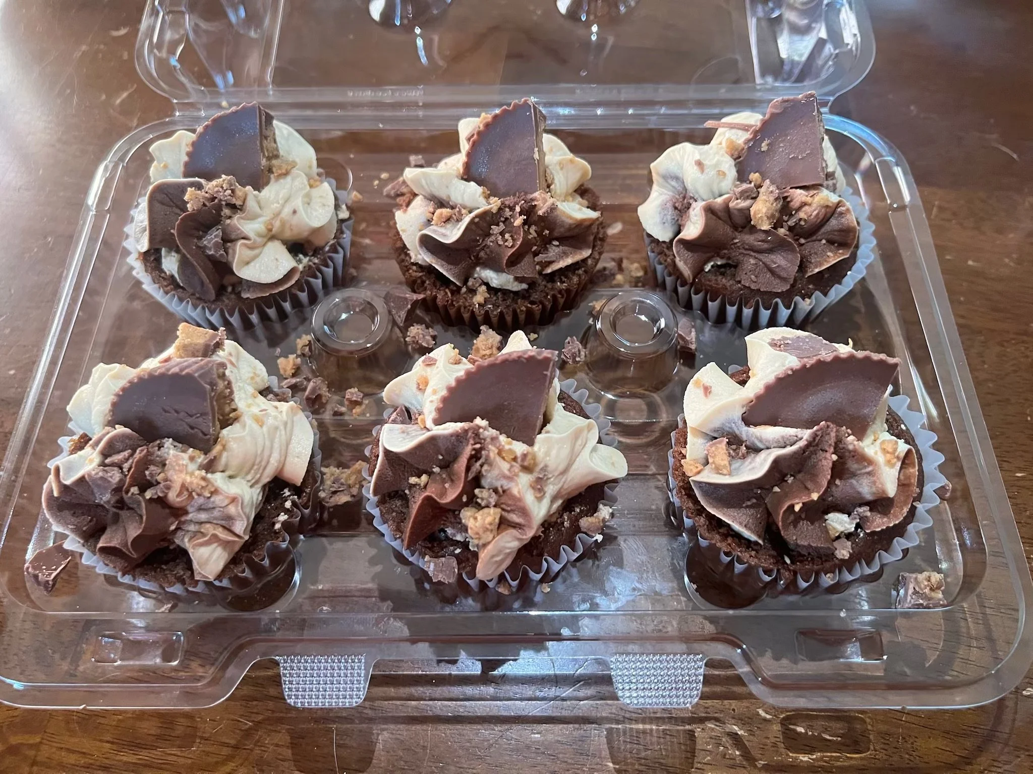 Six chocolate cupcakes decorated with white and dark chocolate swirls and a piece of chocolate on top, placed in a clear plastic container on a wooden surface.