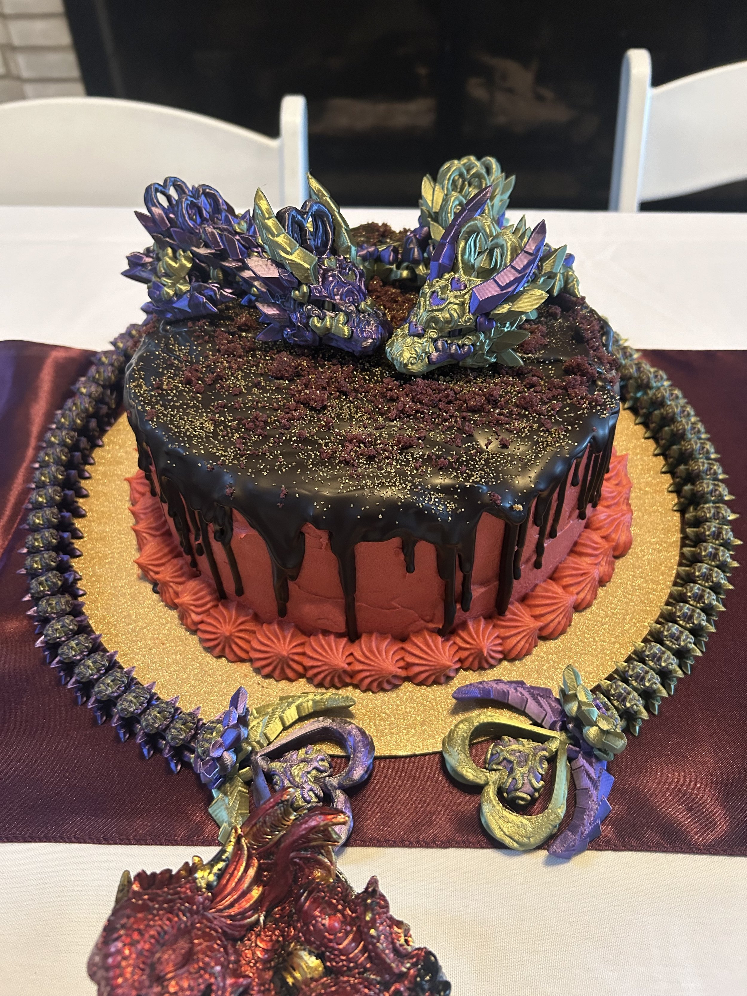 A cake decorated with dragon figures and gold accents, featuring a black top with drip icing, pink frosting on the sides, and a gold base, surrounded by a decorative border with dragon figures.