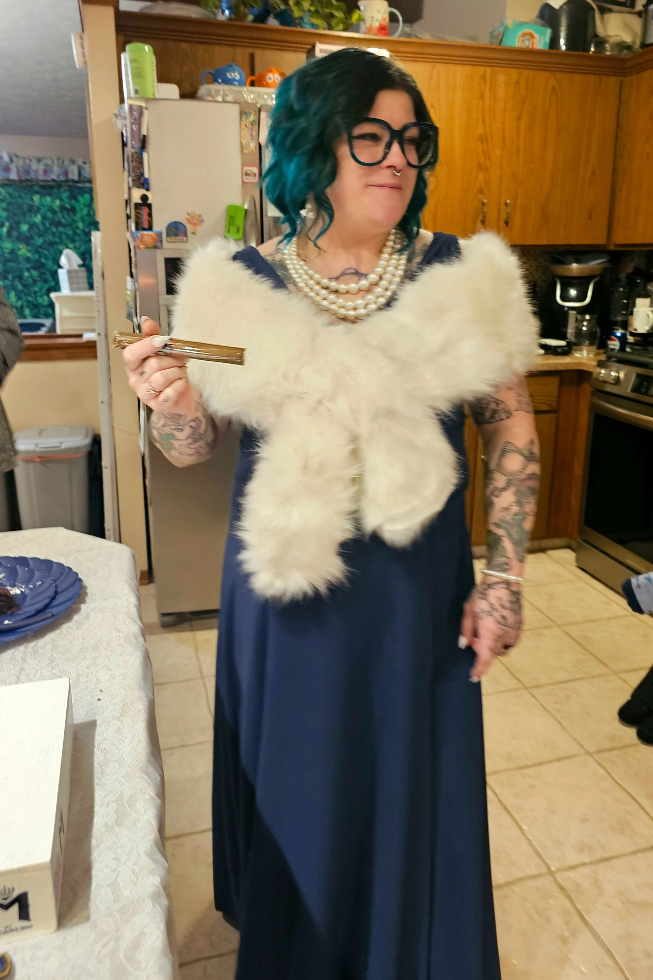 Woman with blue and black hair, wearing glasses, pearl necklaces, and a navy blue dress, holding a gold-colored object, standing in a kitchen with wooden cabinets and various decorative items.