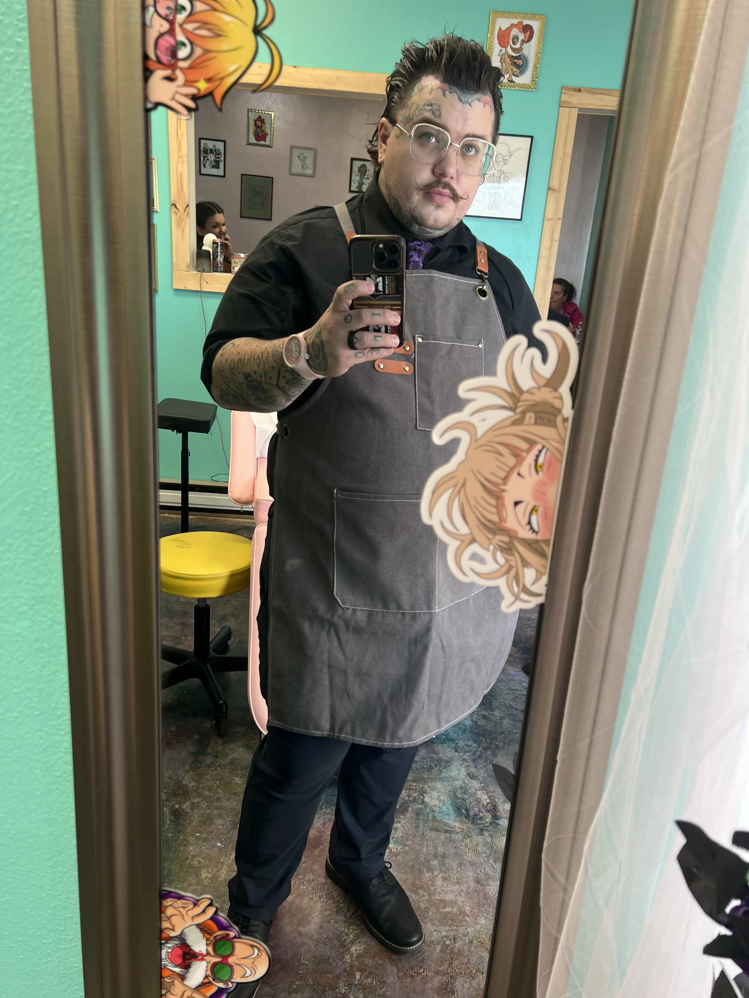 A person with tattoos and glasses taking a mirror selfie in a colorful parlor, wearing a black shirt, gray apron, and black shoes, with cartoon character stickers on the mirror and around the room.