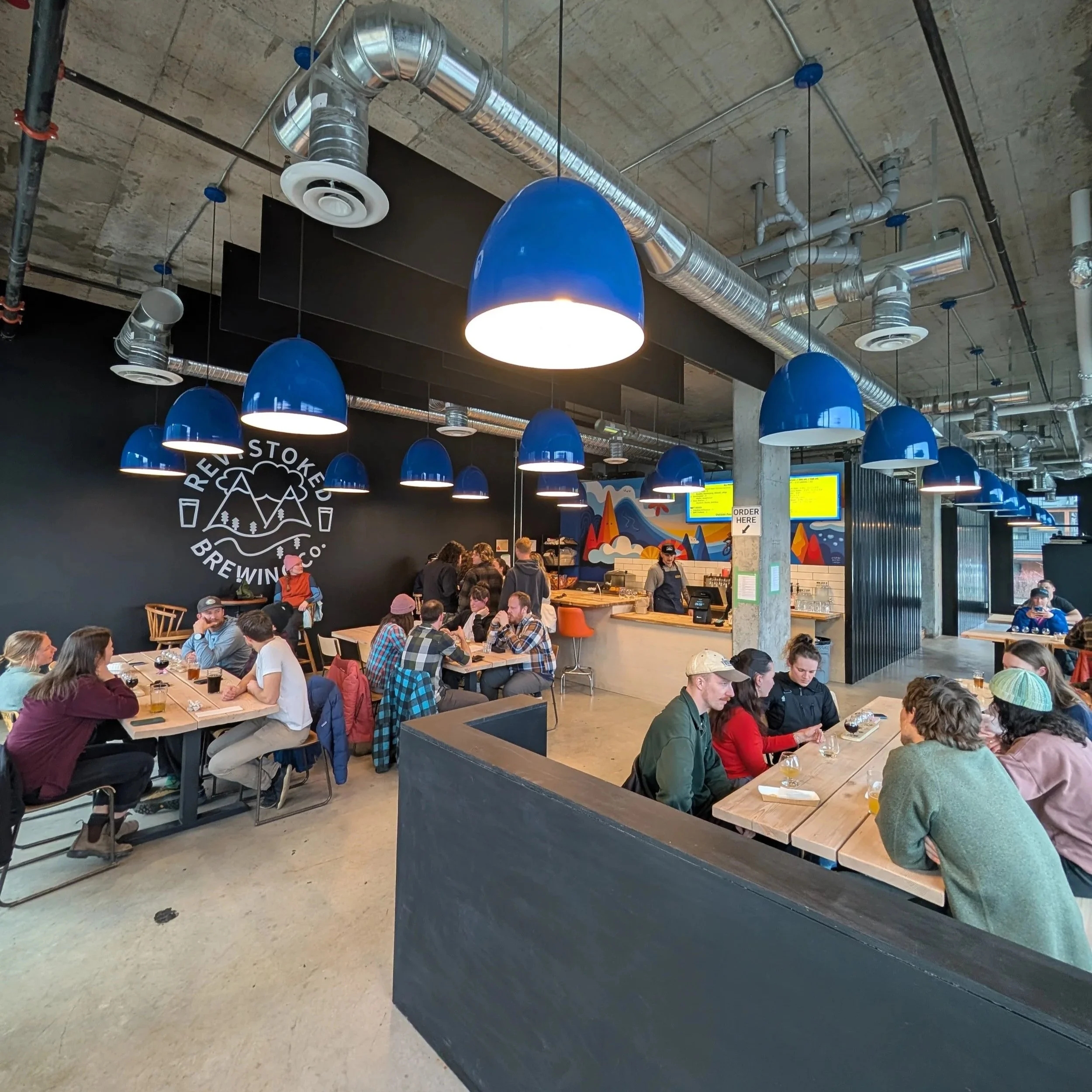 Inside a busy brewery or restaurant, with multiple blue pendant lights hanging from the ceiling, and a wall with the logo of Reuben's Brews Brewing Co. visible. People are seated at tables and the counter is visible in the background.