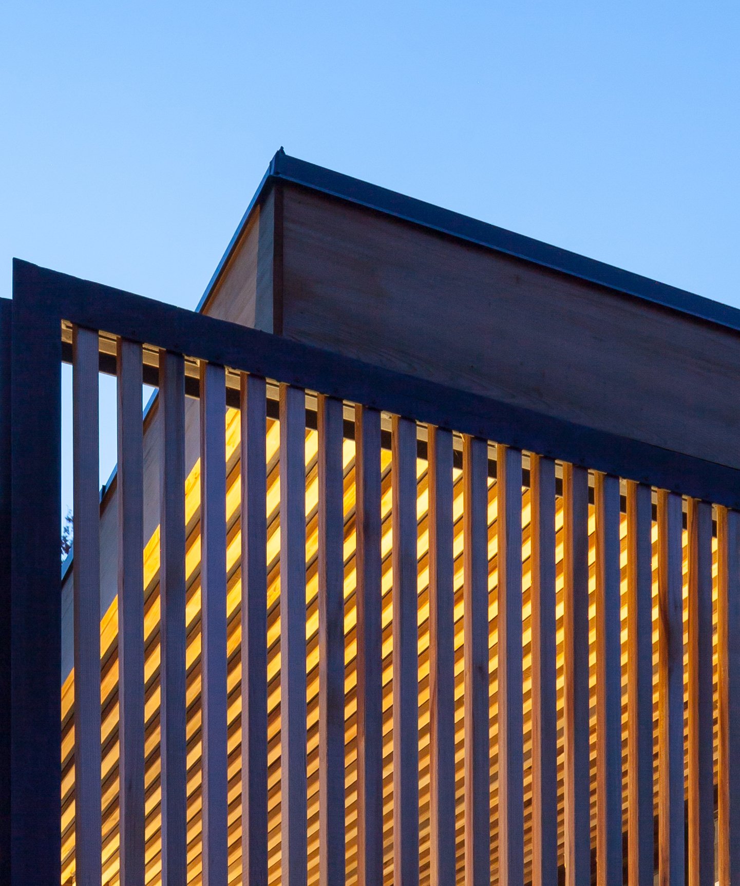 A close-up of a modern architectural screen composed of evenly spaced wooden slats, illuminated by warm interior lighting. The slatted design filters light and shadows, creating a dynamic interplay against the evening sky.