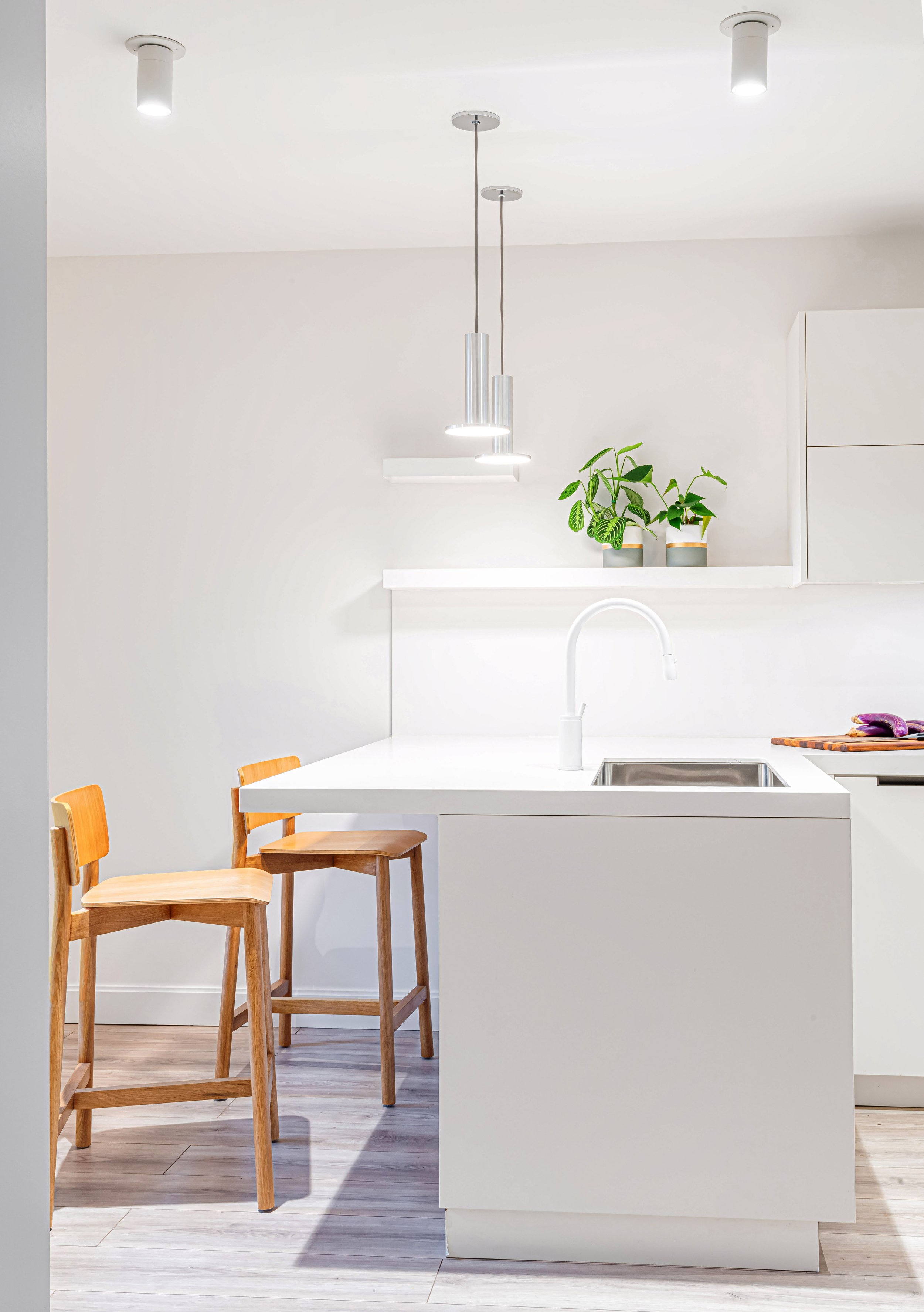 A bright, modern kitchen featuring a minimalist white peninsula, wooden bar stools, and sleek pendant lighting. The space is illuminated by natural light, with integrated storage and a warm material palette, reflecting Poiesis Architecture’s approach