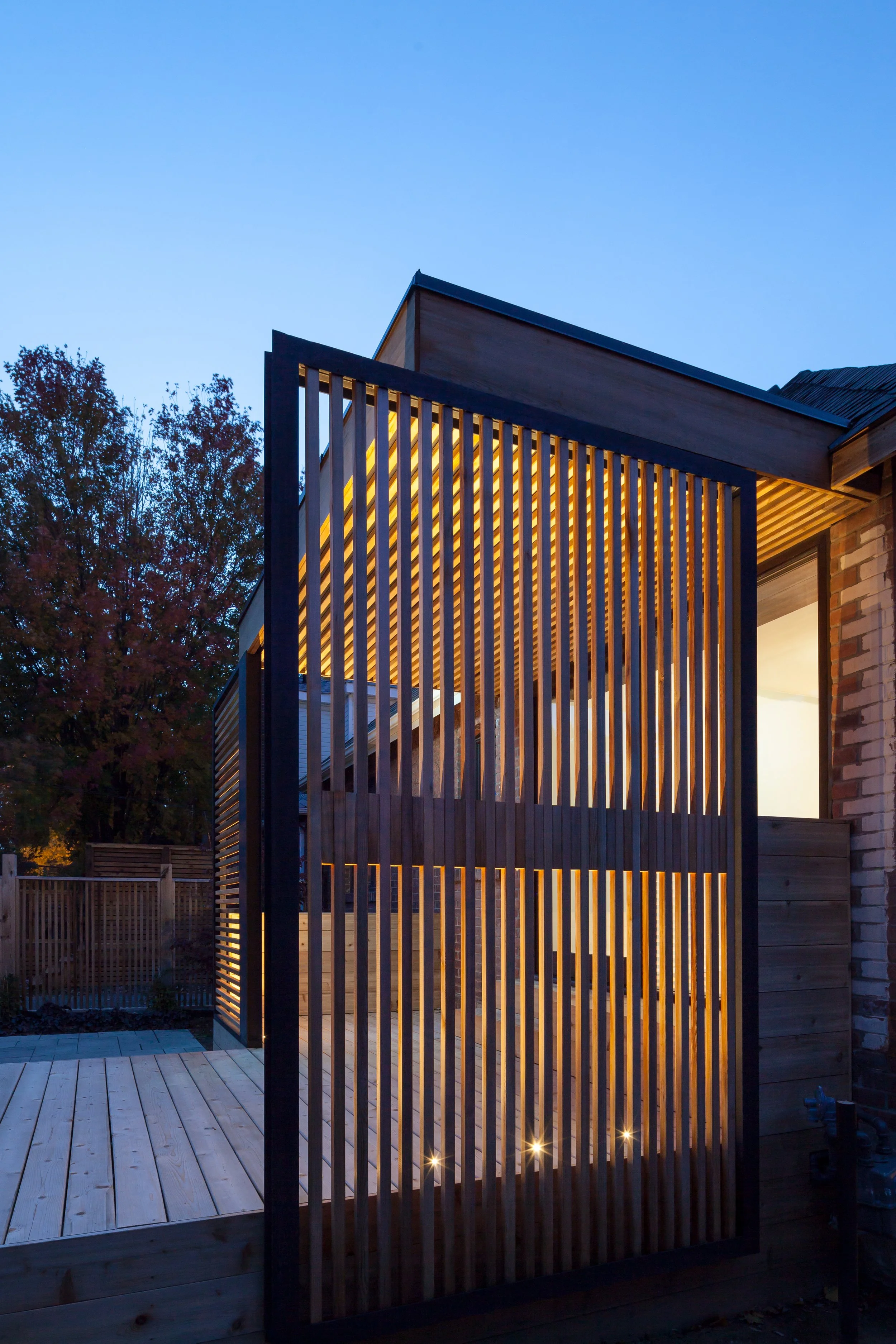A contemporary wood terrace illuminated at dusk, with slatted wood screens framed in black metal. The dynamic lighting highlights the texture and craftsmanship of the outdoor space, showcasing Poiesis Architecture’s attention to materiality and spati