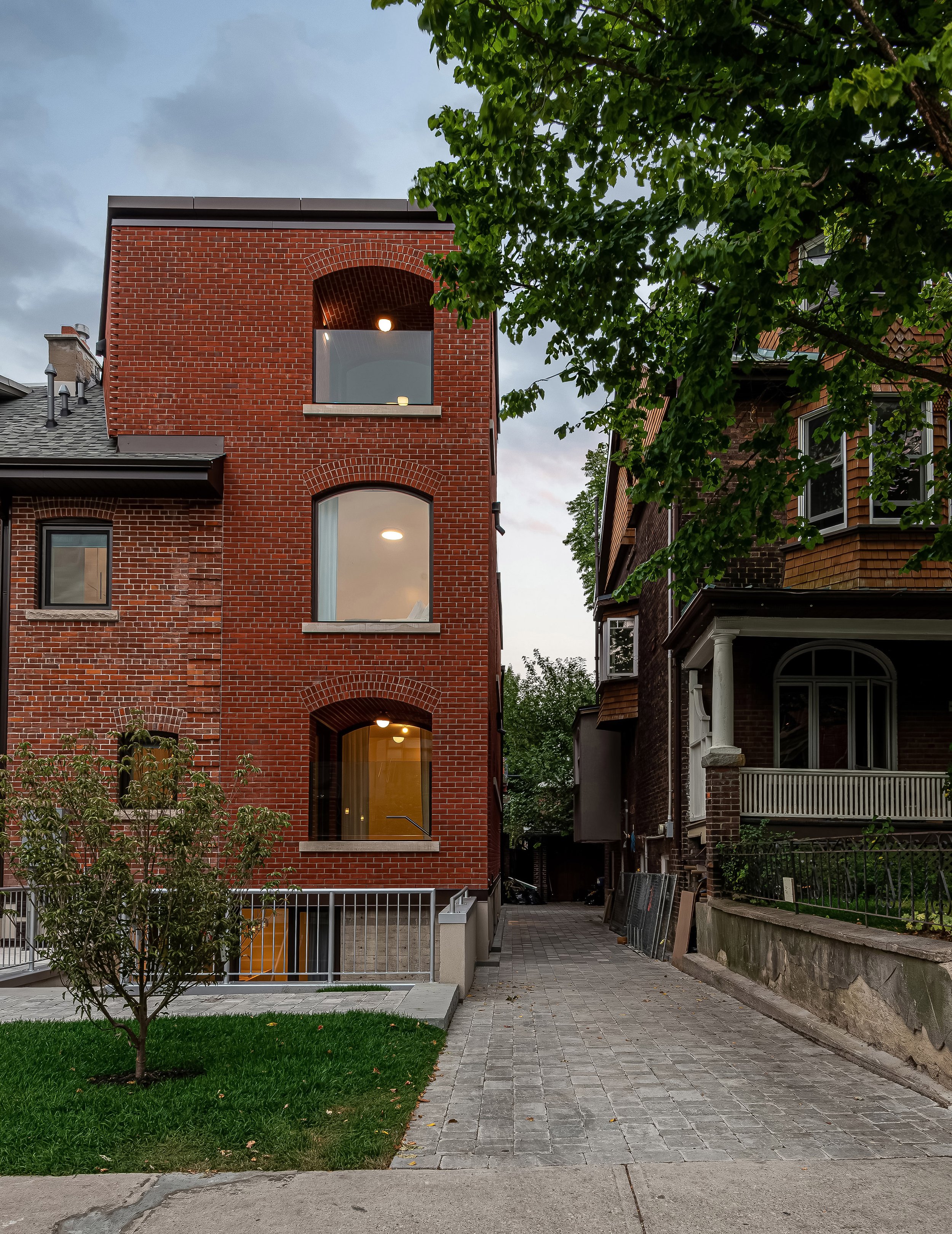 A contemporary red brick fourplex featuring arched windows, warm interior lighting, and a landscaped entryway. The design blends heritage-inspired materials with modern residential architecture.