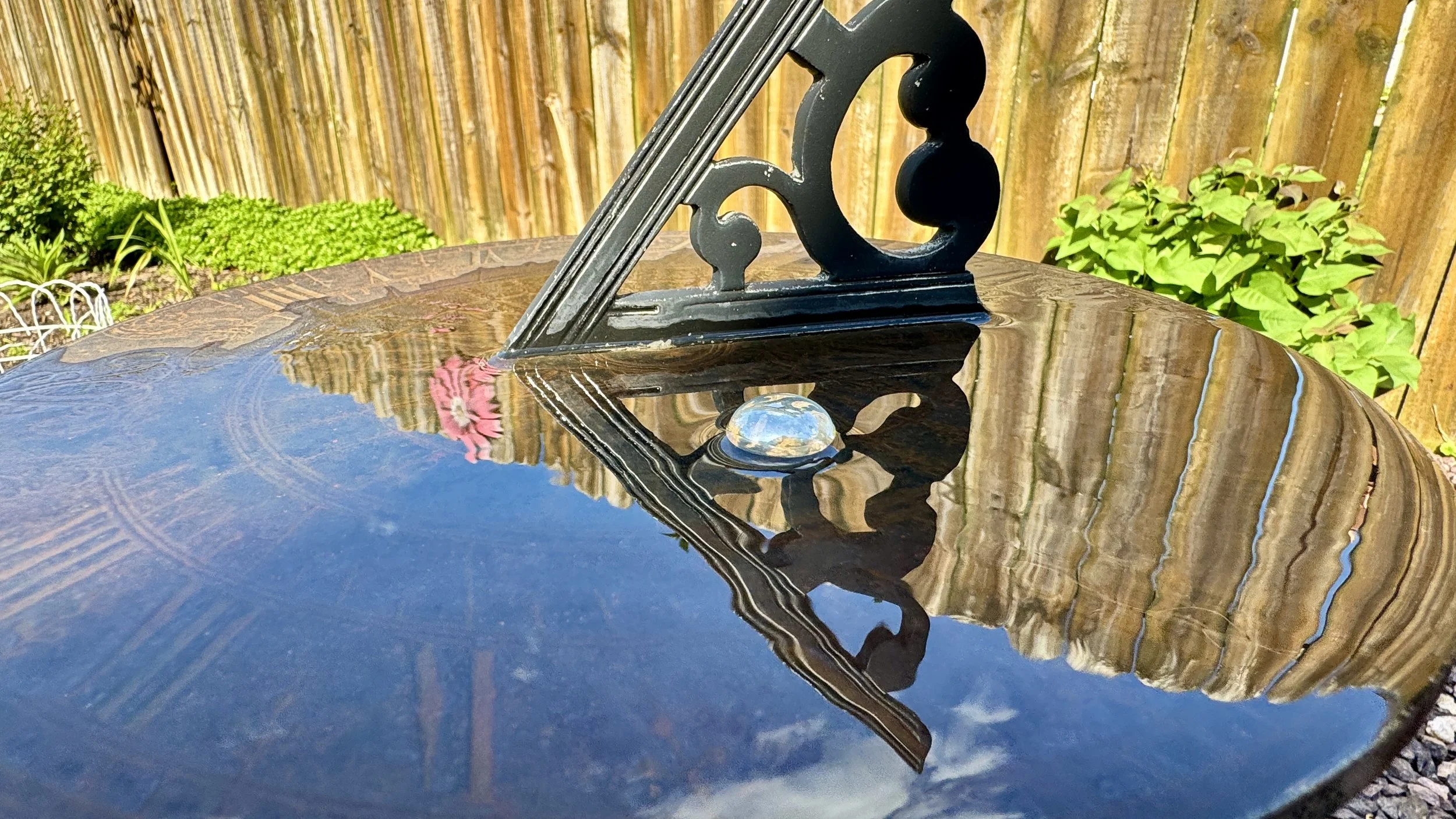 A metal square and triangle tool is placed on a wet, reflective black surface, with a wooden fence and greenery in the background.