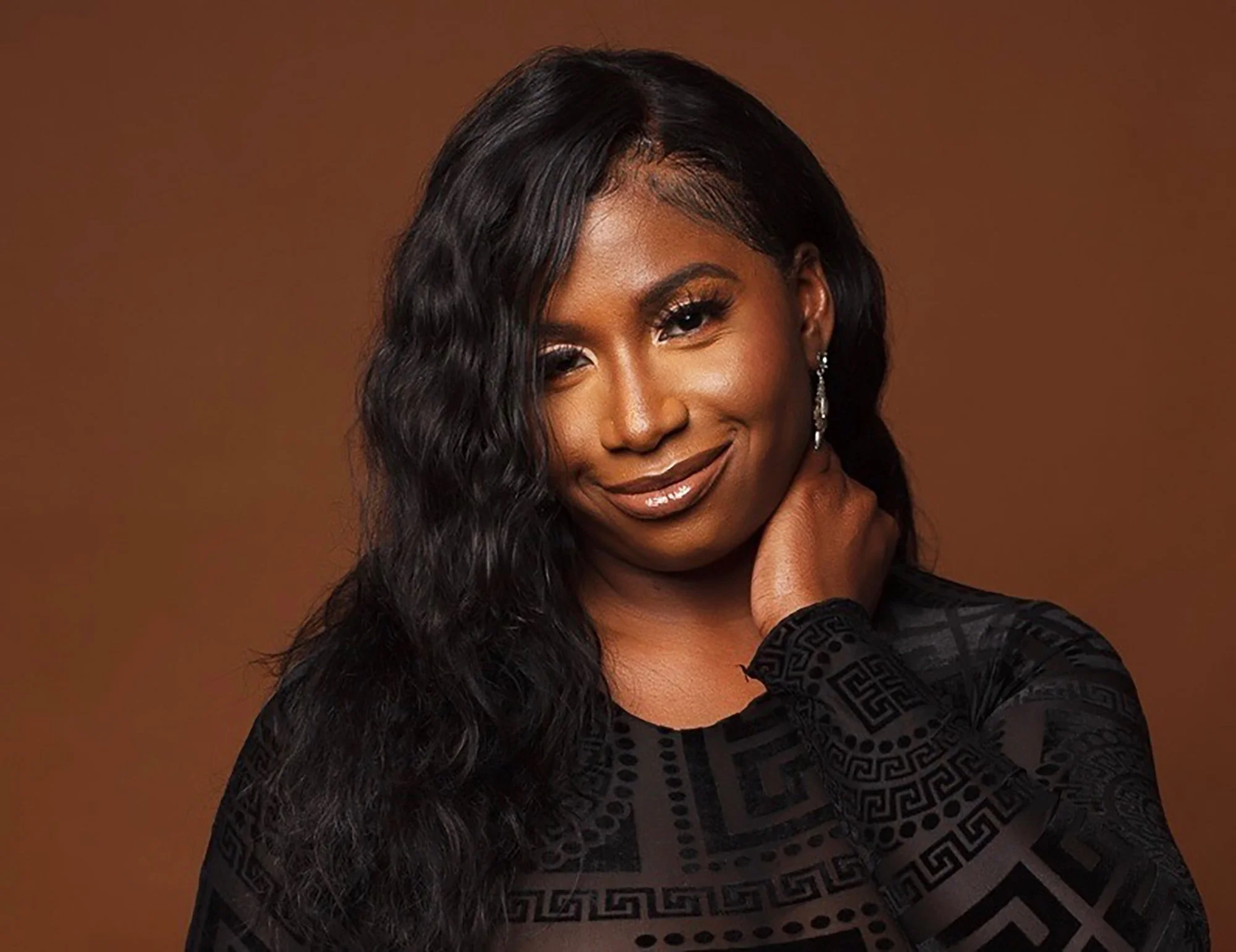 Portrait of an African American woman with long wavy black hair and earrings, wearing a black patterned top, smiling gently on a brown background.