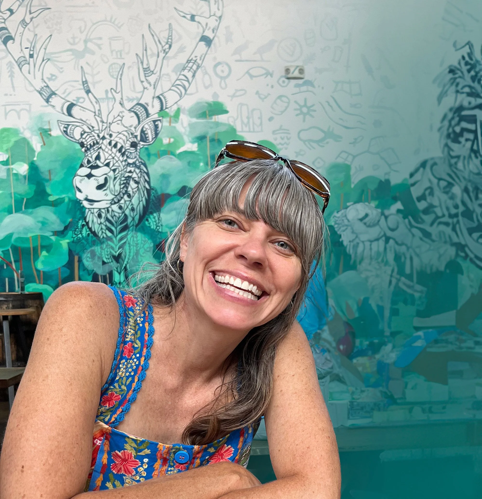 Close-up of a woman smiling with glasses on her head, wearing a sleeveless dress with a colorful floral pattern, positioned in front of a mural featuring a stylized tiger with intricate black and white designs, green foliage, and abstract doodles.