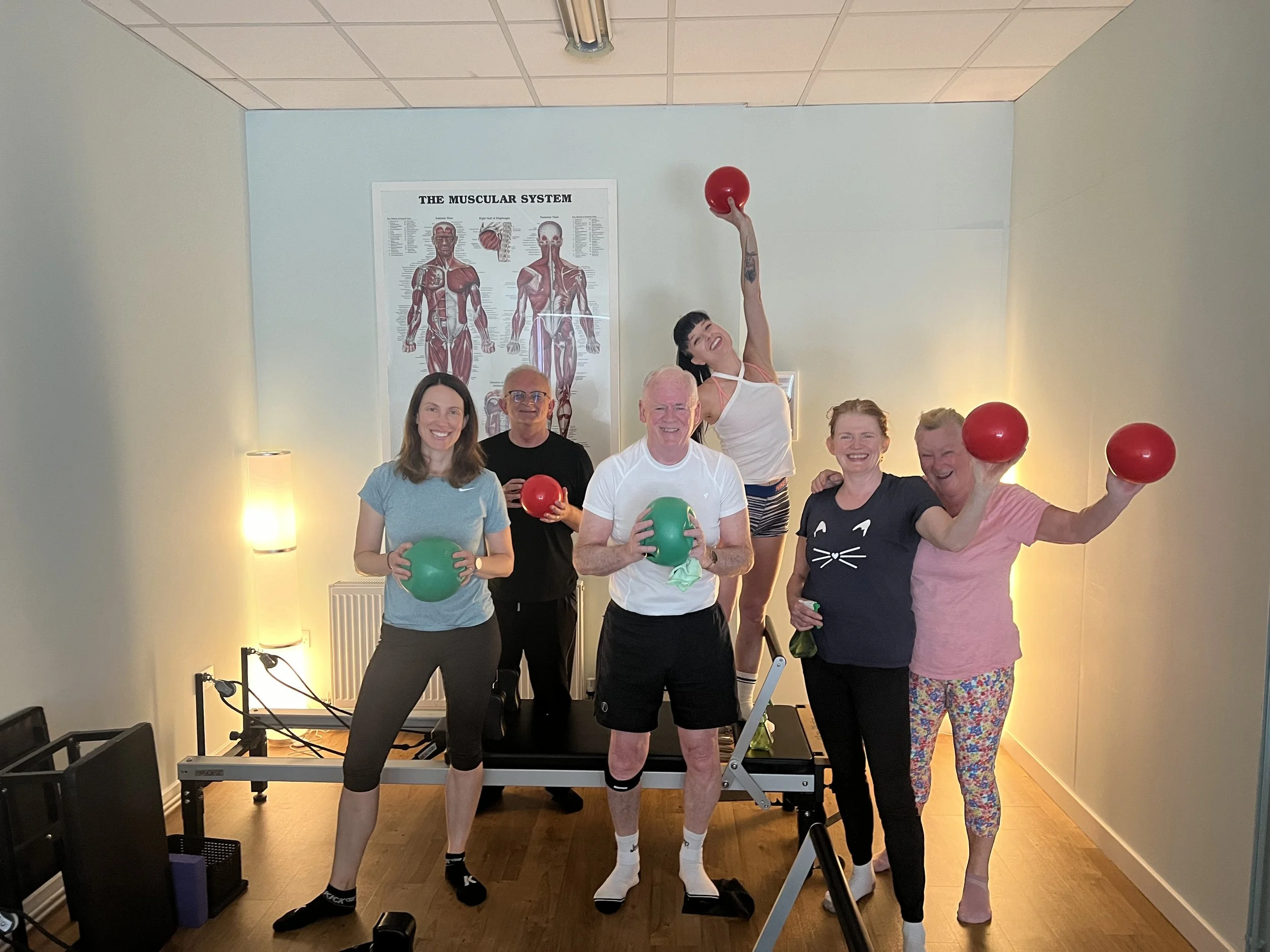 A group of six people in workout attire standing indoors, holding exercise balls. They are posing in a fitness studio with a poster of the muscular system on the wall behind them. There is a Pilates reformer in the foreground and warm lighting in the