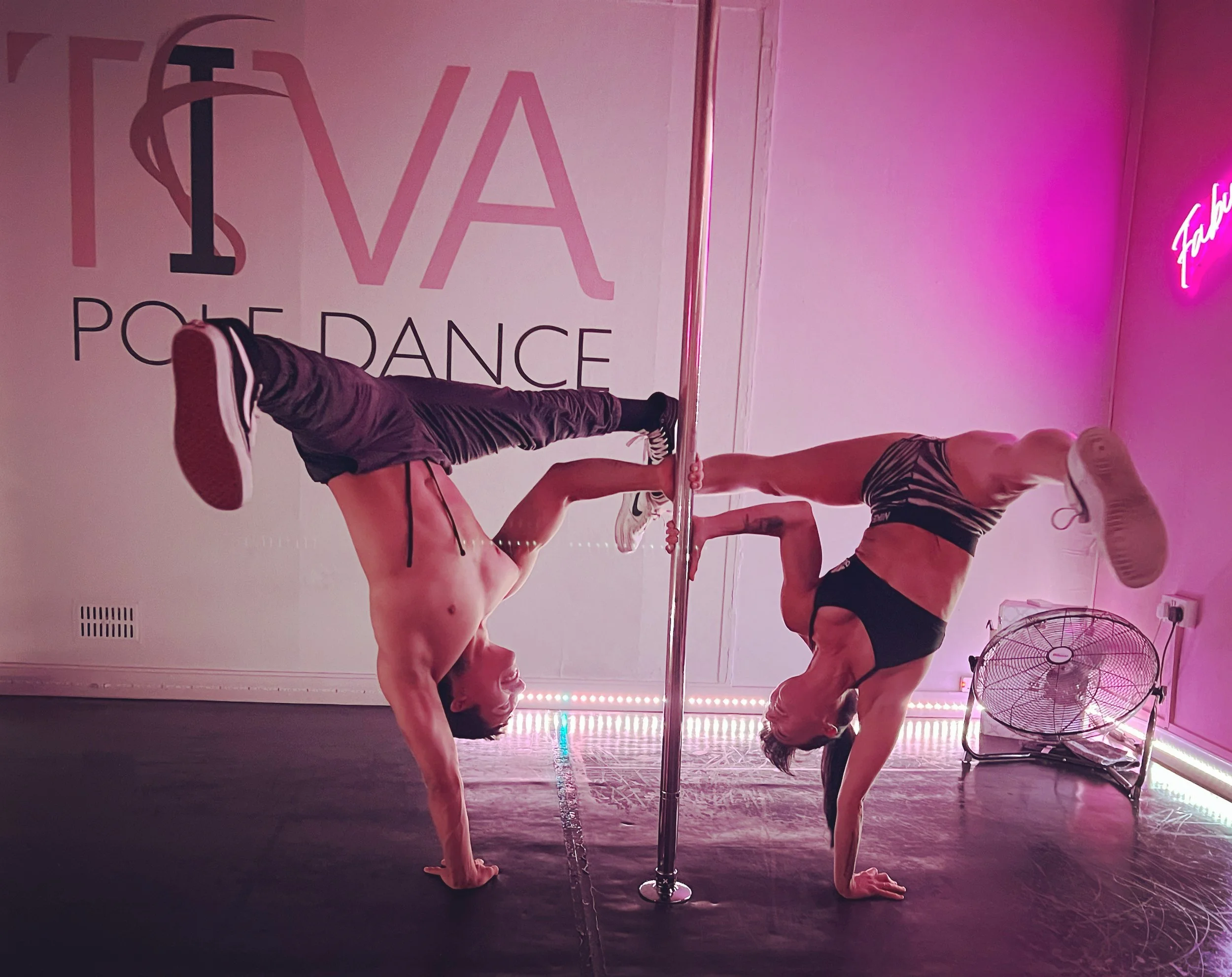Two women performing a pole dance routine, each balanced on one hand with their legs extended and gripping the pole, in a pink-lit studio.