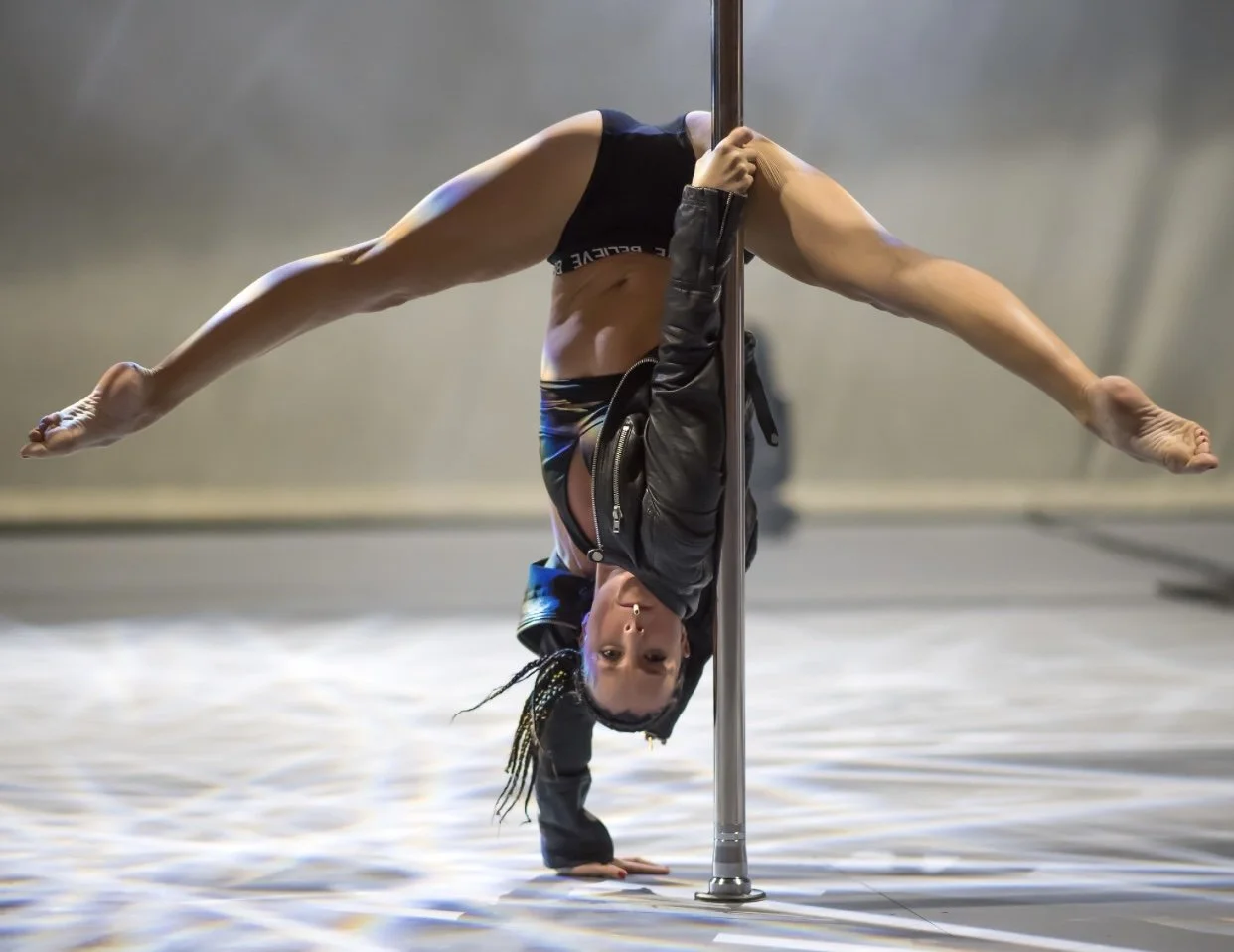 A pole dancer performing an acrobatic move, hanging upside down with legs extended, wearing a black jacket and shorts.