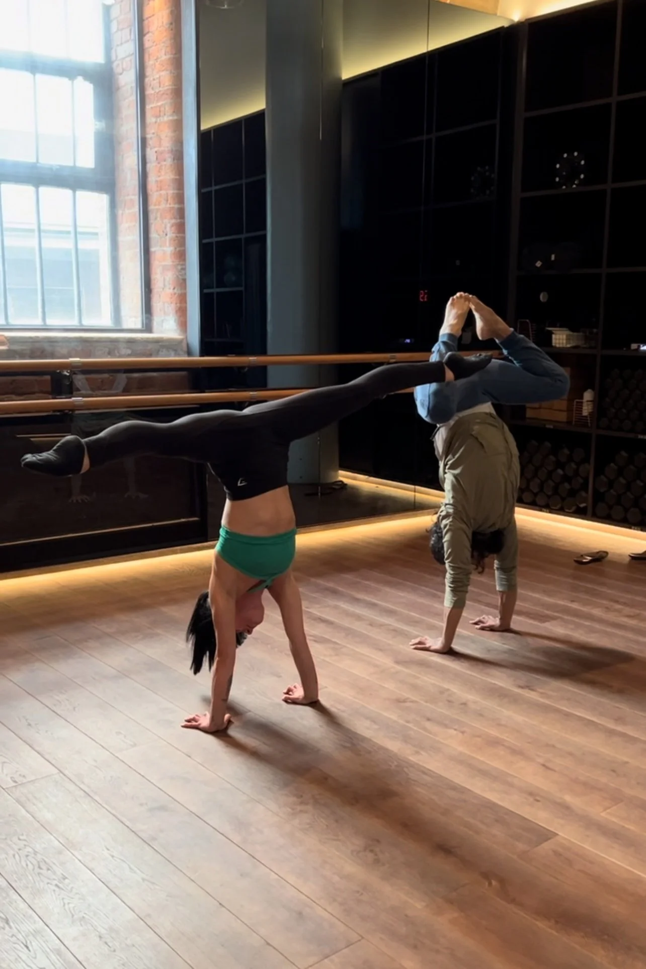 Two people practicing acro yoga in a dance studio, one person balancing on their hands while supporting the other person who's upside down with their legs extended horizontally.