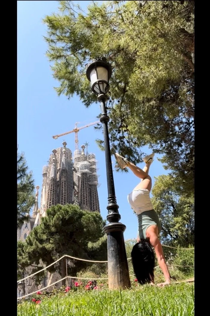 Woman in athletic wear performing a handstand outdoors near a lamppost, with the Sagrada Familia cathedral under construction visible in the background in Barcelona.