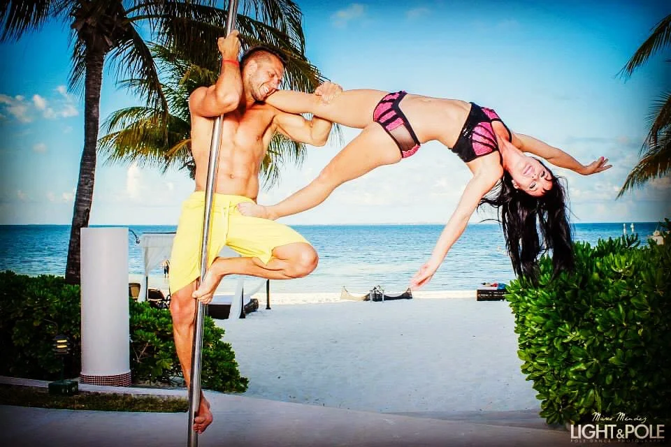 A man practicing pole dance with a woman outdoors on a beach, with palm trees and ocean in the background.