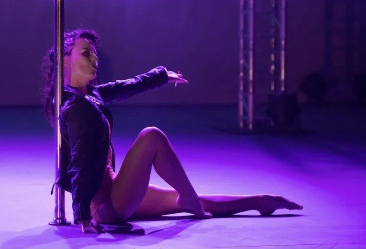 A person performing a pole dance move on stage with purple lighting.