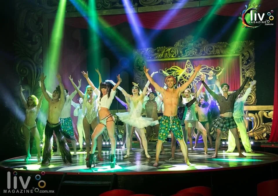 A group of performers on stage with colorful lighting, wearing diverse costumes, posing with arms raised. The background features ornate decorations and a red curtain.