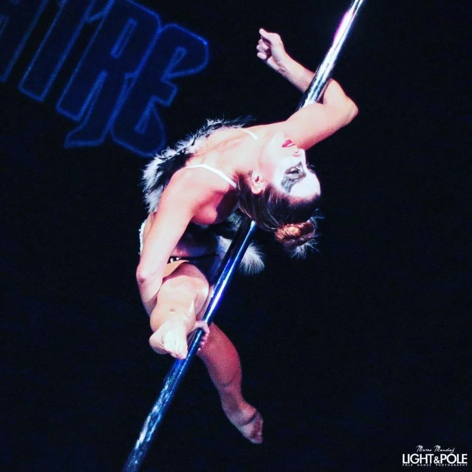 Pole dancer performing a pose on stage, wearing costume and face paint, with dark background.