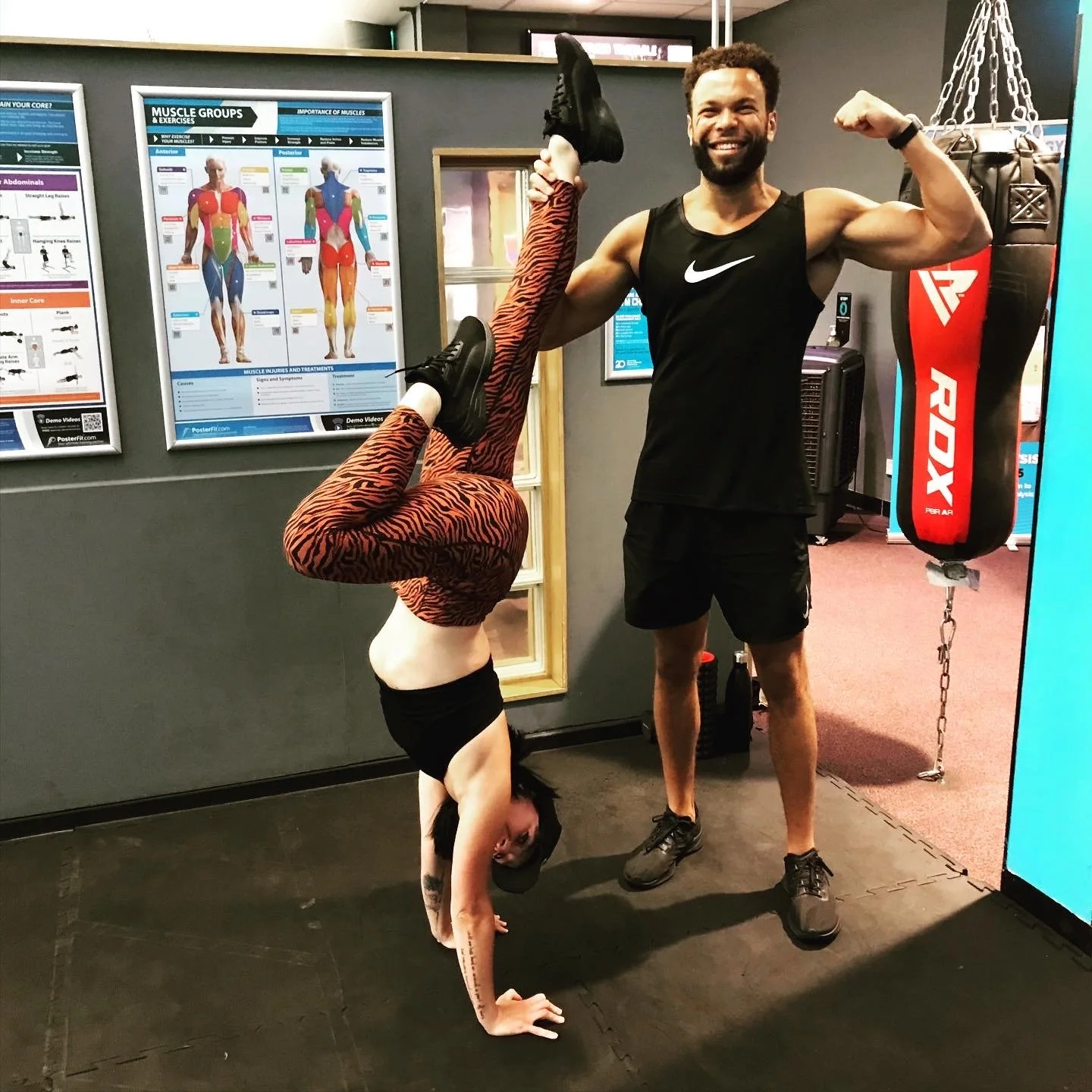 A woman performing a headstand with her legs bent at the knees and hands on the ground, wearing tiger print leggings and black top, in a gym. A man is standing beside her, smiling and flexing his arm, wearing a black sleeveless Nike shirt and black s