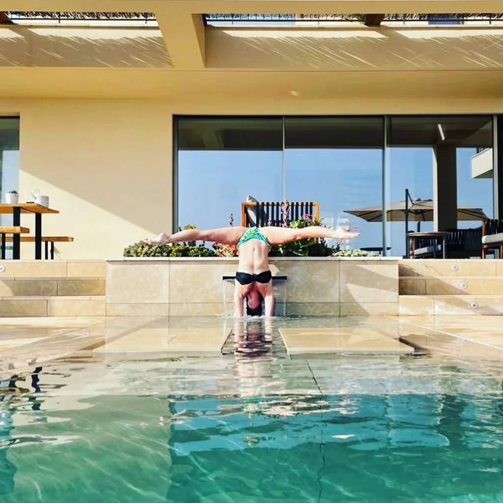 Person doing a handstand split over a pool, with a modern house and patio in the background.
