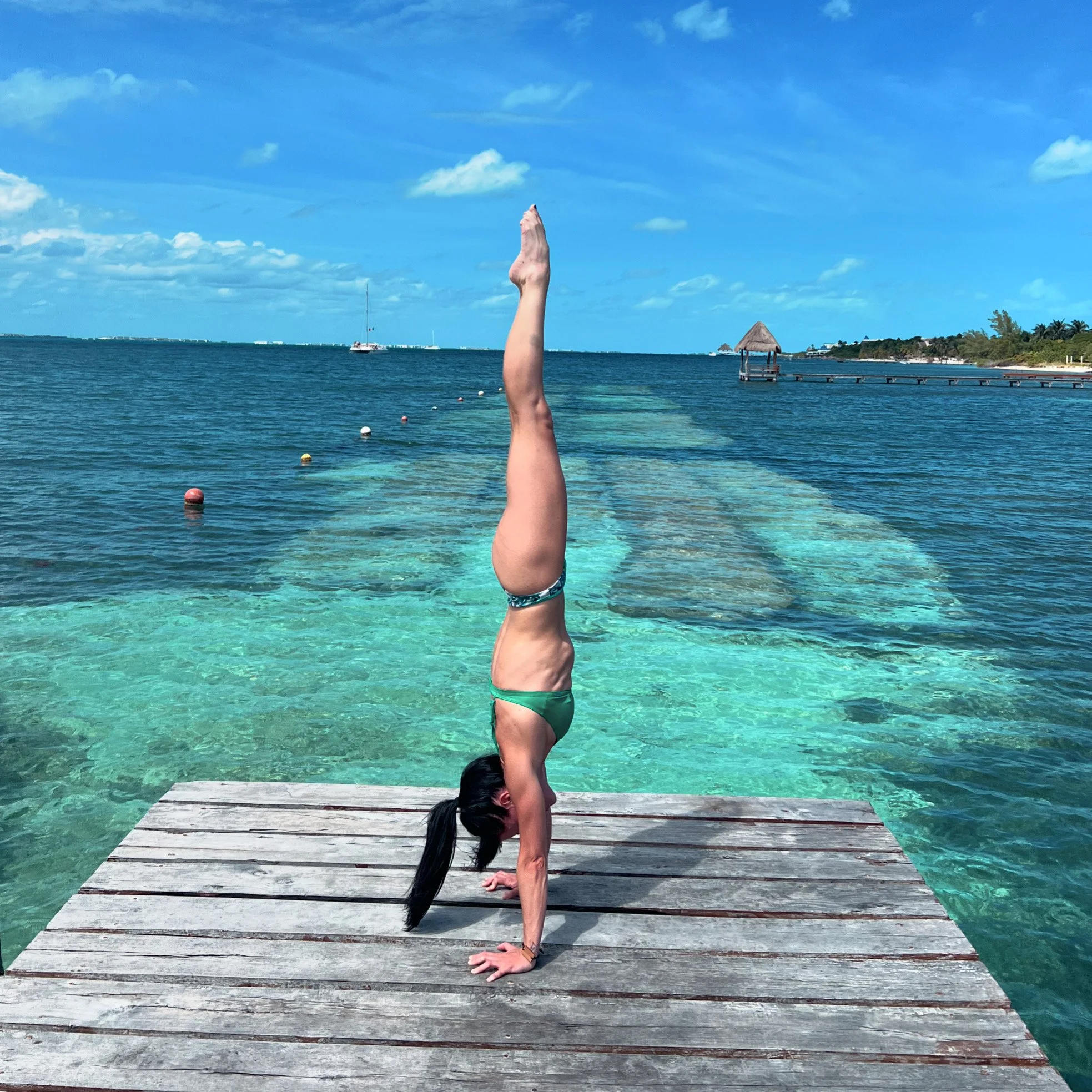 Person in green bikini performing a handstand on a wooden pier by the ocean with clear blue water and a sunny sky.