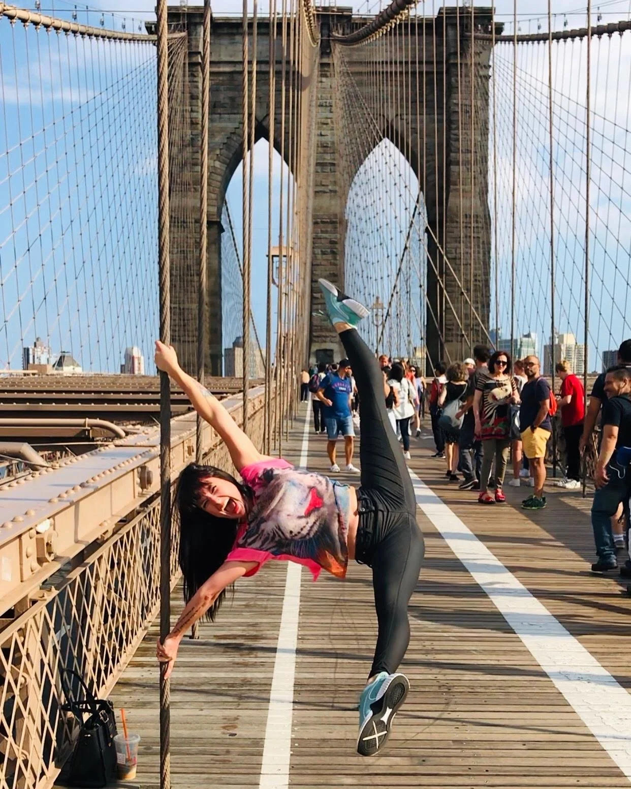 A woman in colorful clothes striking a pose on the Brooklyn Bridge, with many people walking in the background.