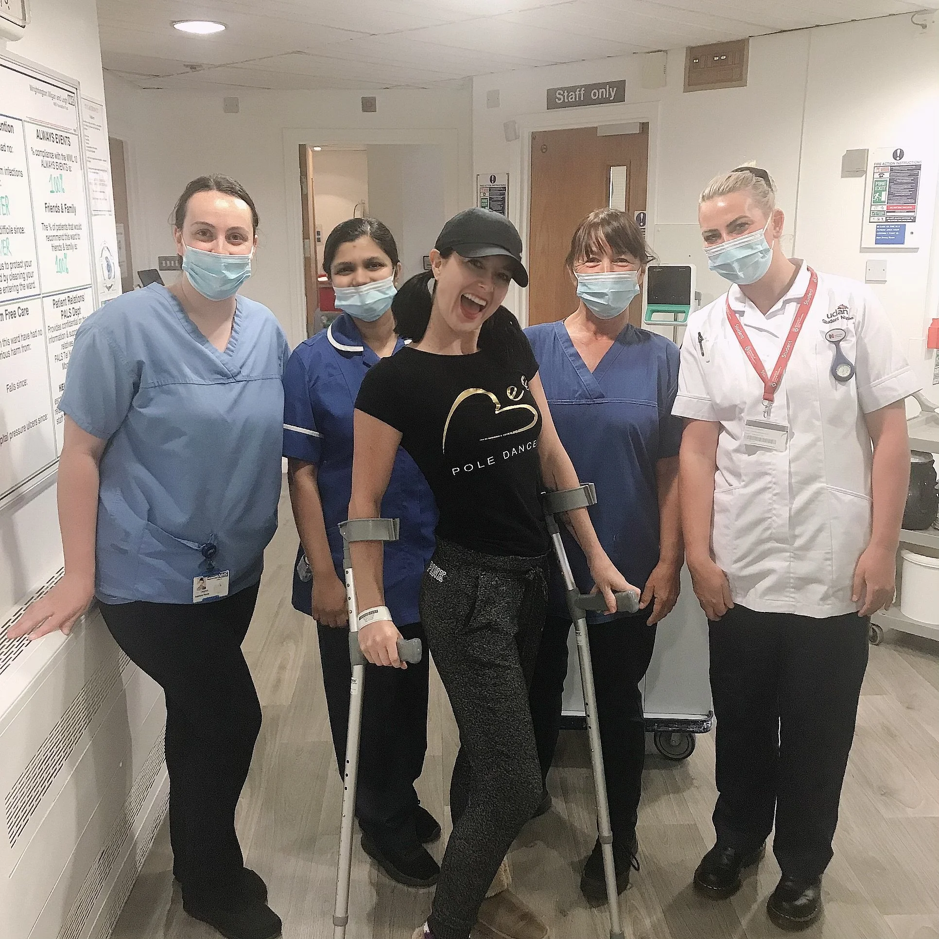 A group of five women, including healthcare workers and a woman on crutches, standing together in a hospital corridor, all wearing face masks and smiling.