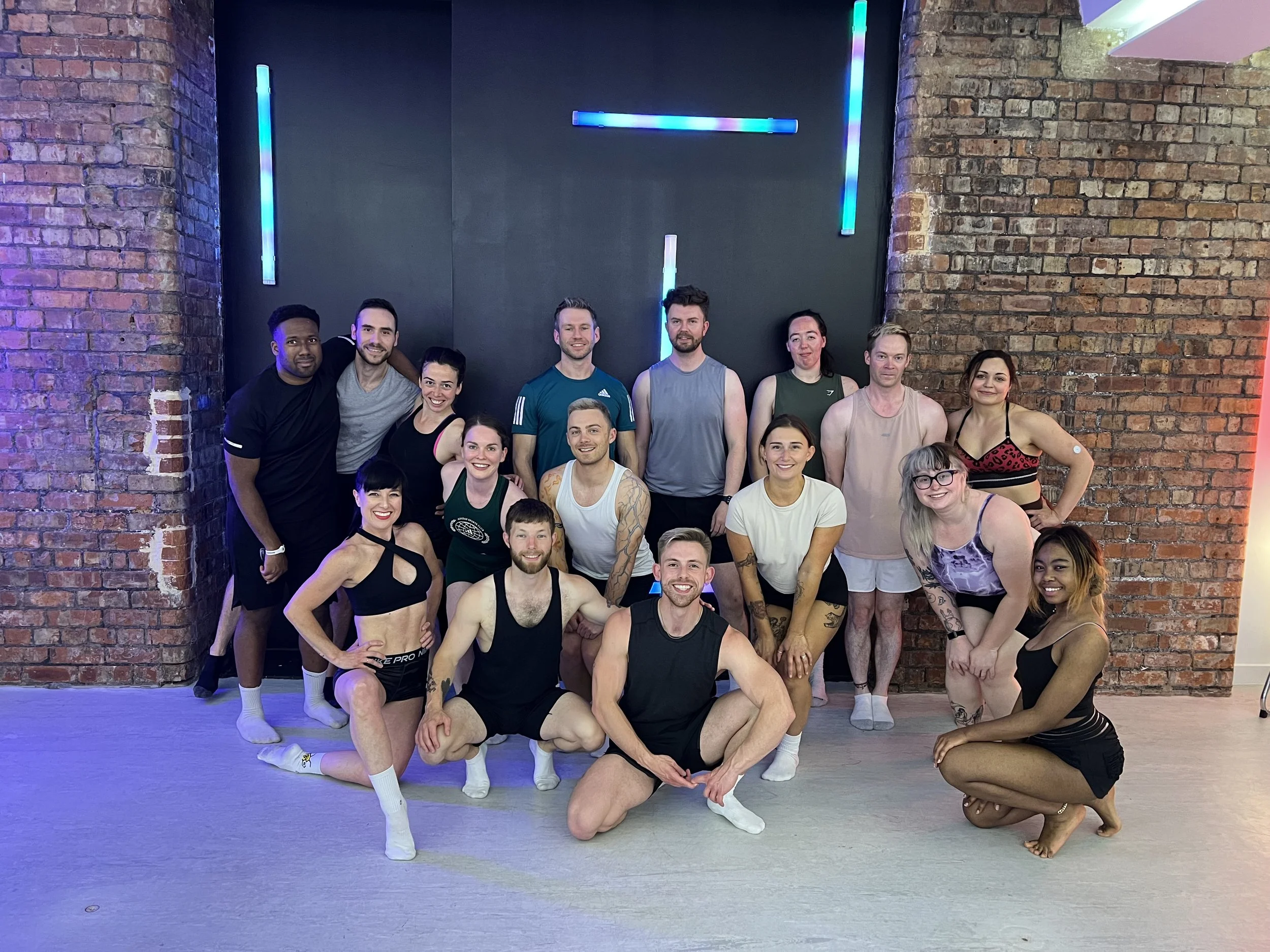 A group of diverse dancers in athletic wear posing together against a brick wall with neon lights.