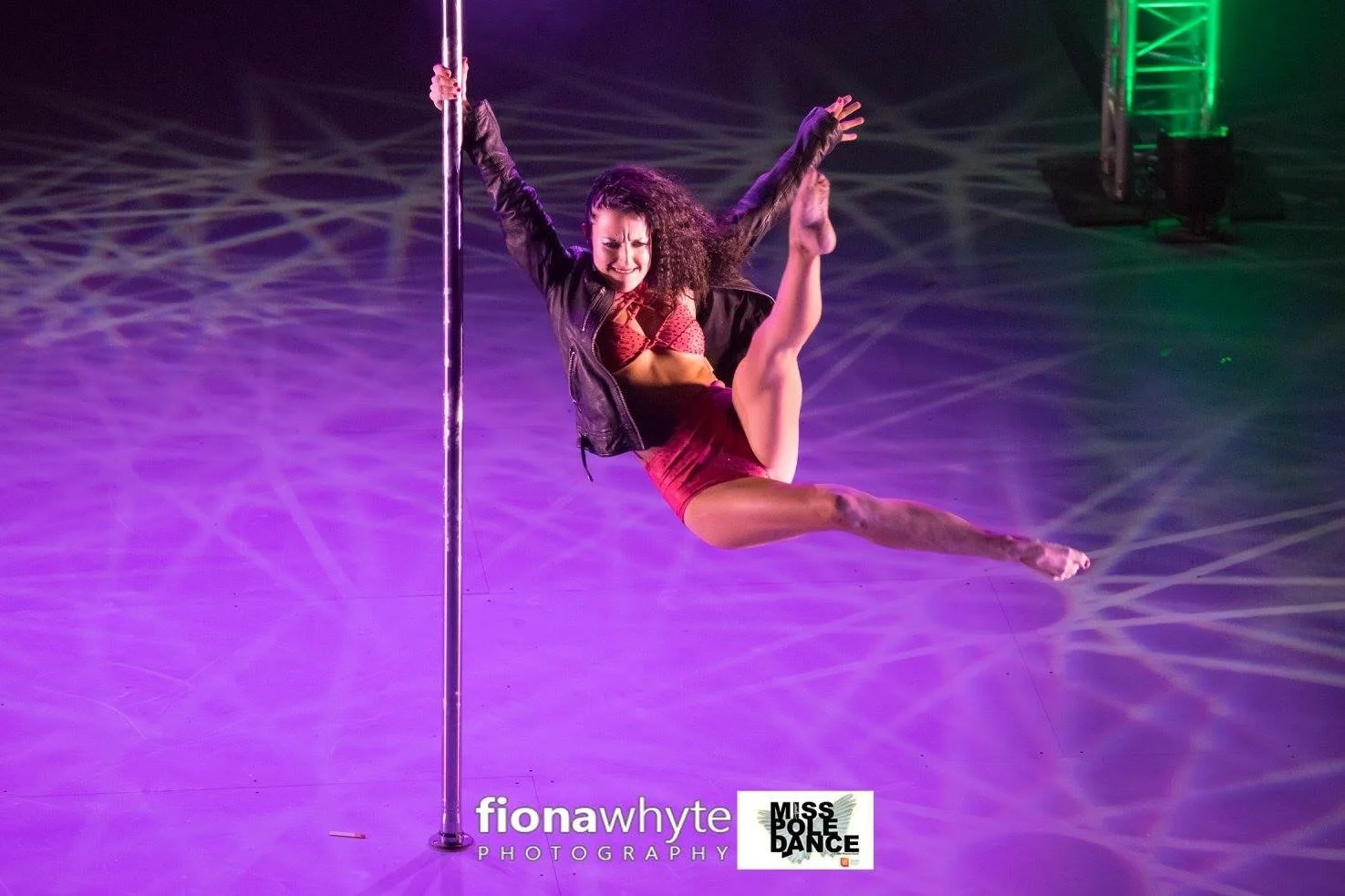 A woman performs a pole dance on stage, holding onto the pole with one hand, with her body extended in a split position, smiling, wearing a red bra and shorts, black jacket, and stage lighting creating colorful effects.