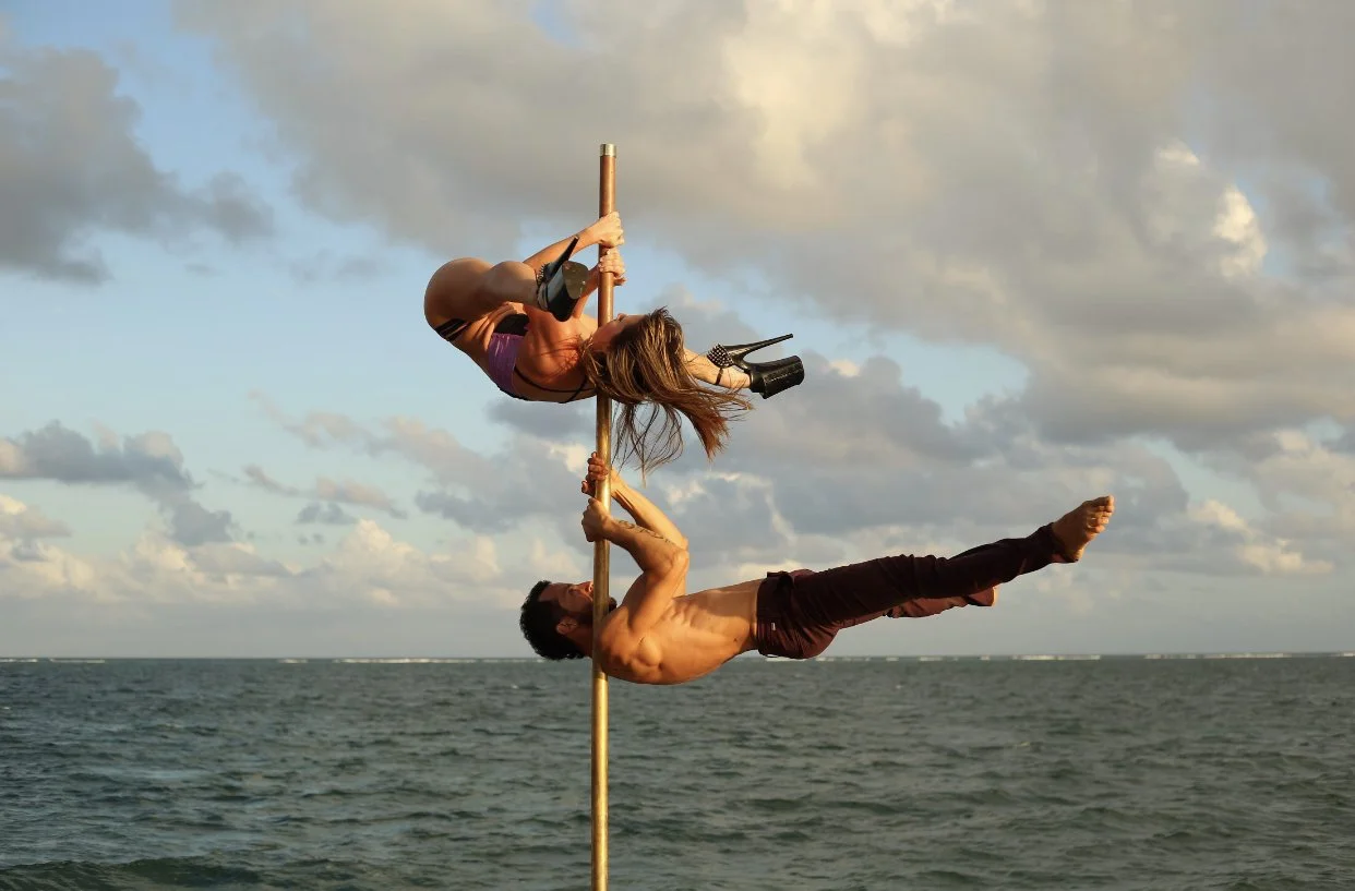 Two performers, a man and a woman, are pole dancing on a pole over the water during sunset. The woman is hanging upside down in a pose, wearing high heels and a purple outfit, while the man supports her from below with his feet extended horizontally.