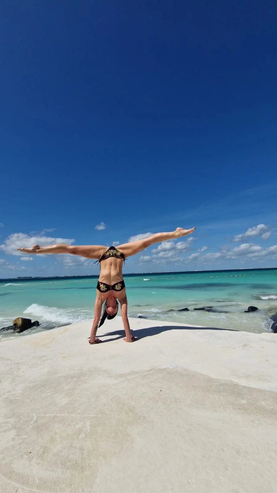 Person in a bikini doing a handstand with legs spread wide on a beach, with turquoise ocean and blue sky in the background.