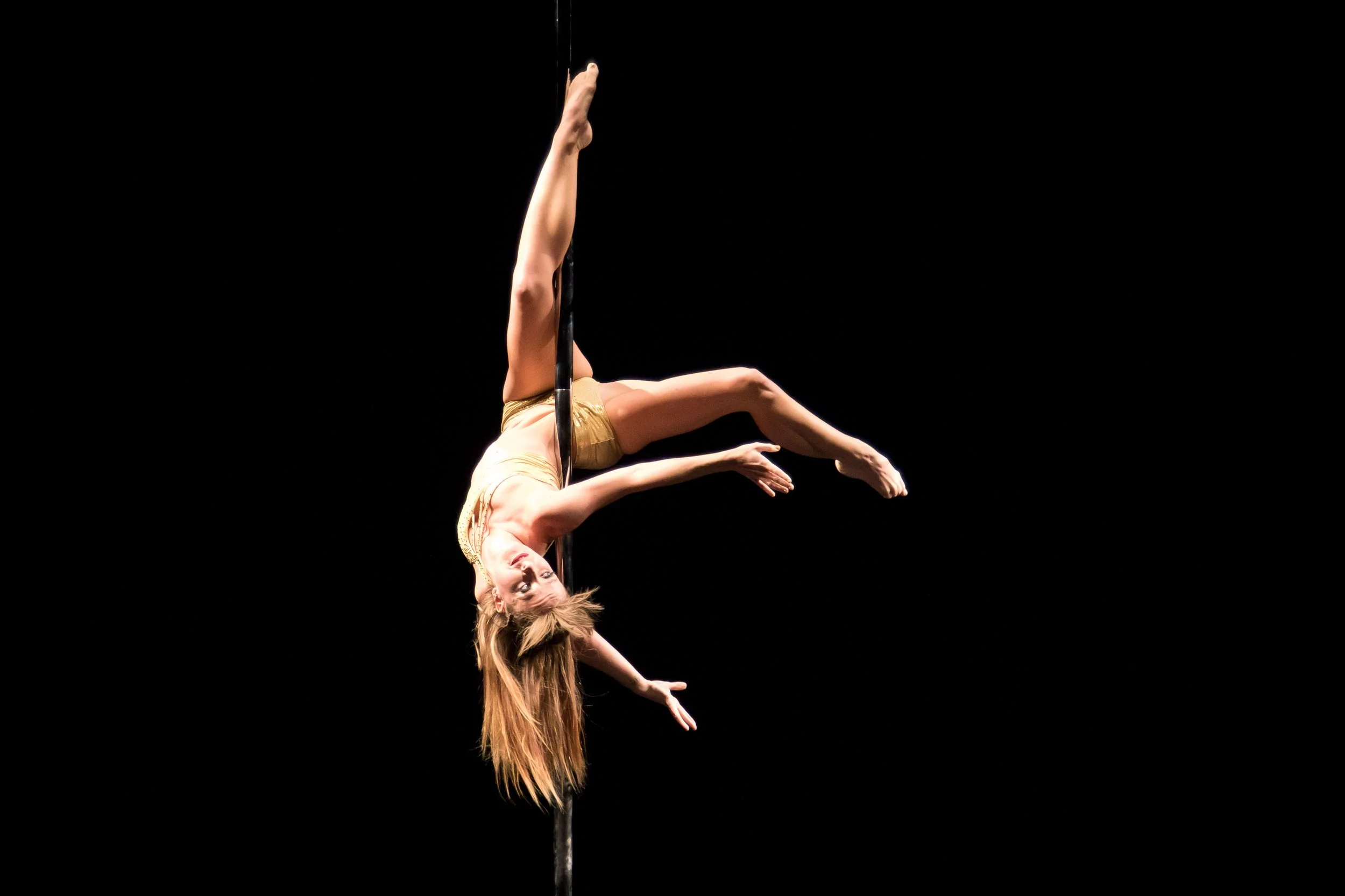 A pole dancer performing an acrobatic move on a pole, wearing a gold outfit, set against a black background.