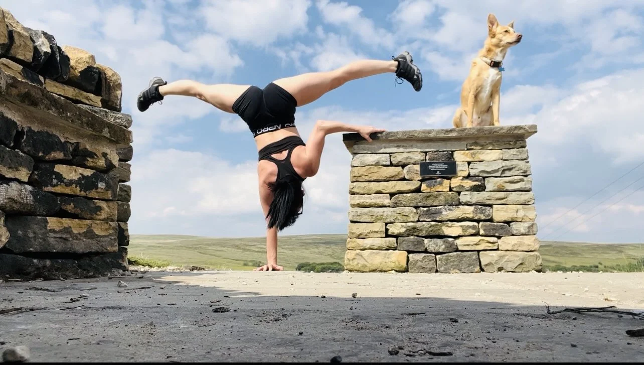 A woman performs a handstand in front of a stone pedestal with a dog sitting on top. The woman is in a one-handed handstand position, and the dog, a small tan mixed breed, is sitting looking off into the distance. The scene is outdoors on a sunny day
