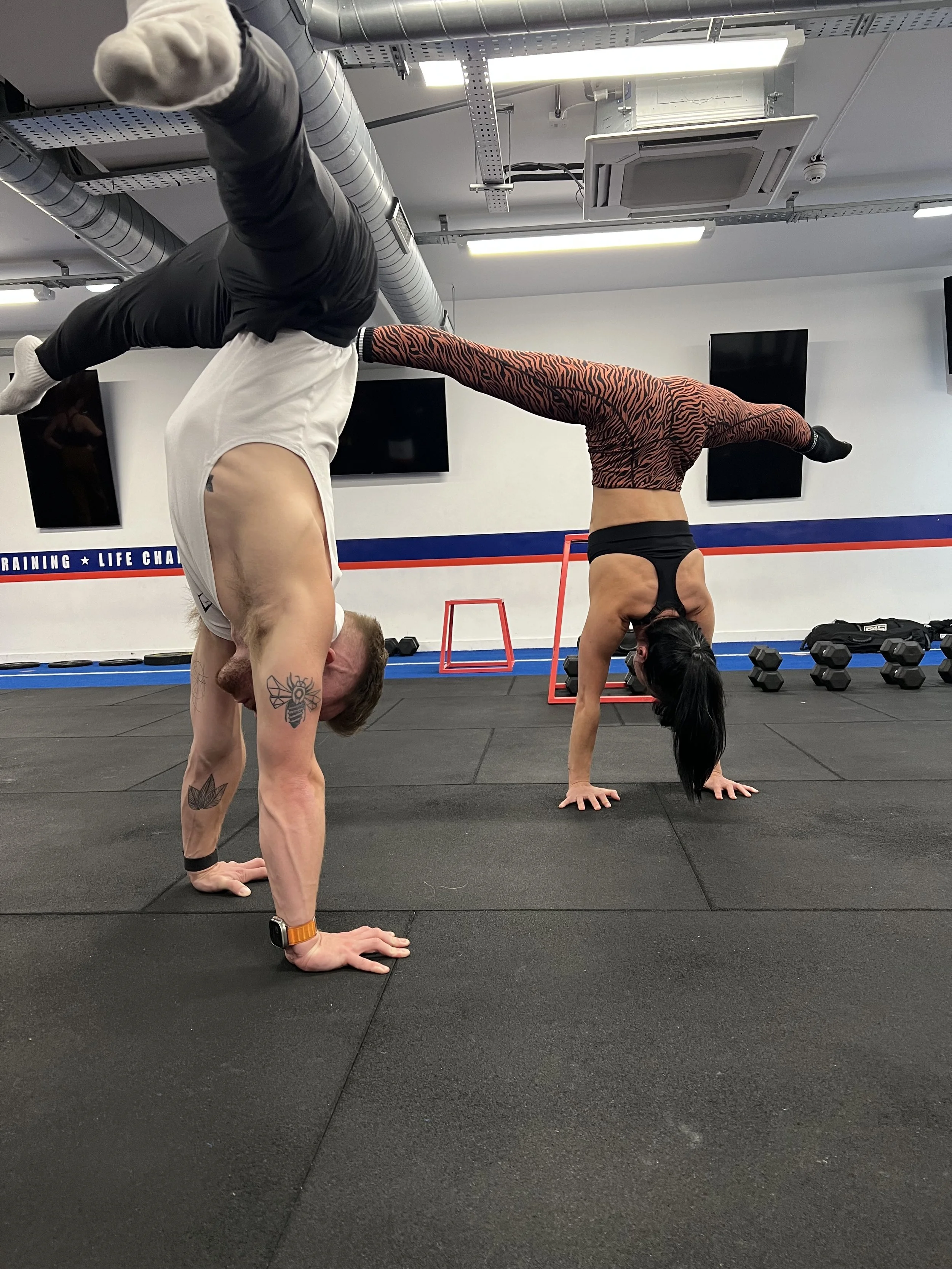 Two people performing a handstand in a gym with weightlifting equipment in the background.