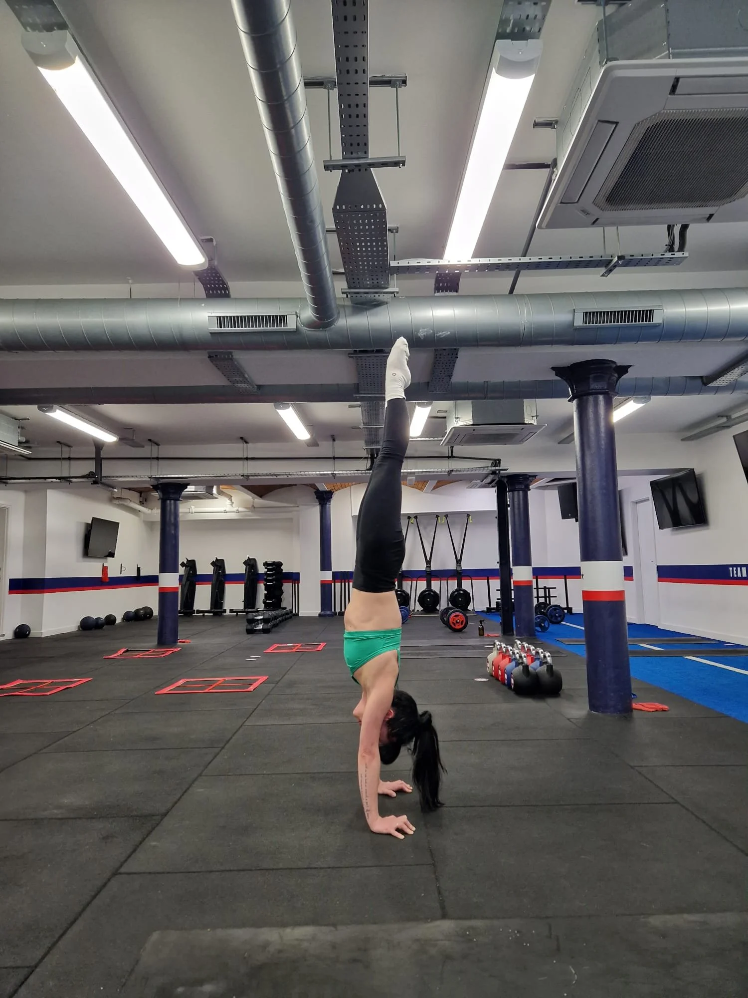 A woman performing a handstand in a gym, with one leg extended straight up towards the ceiling.