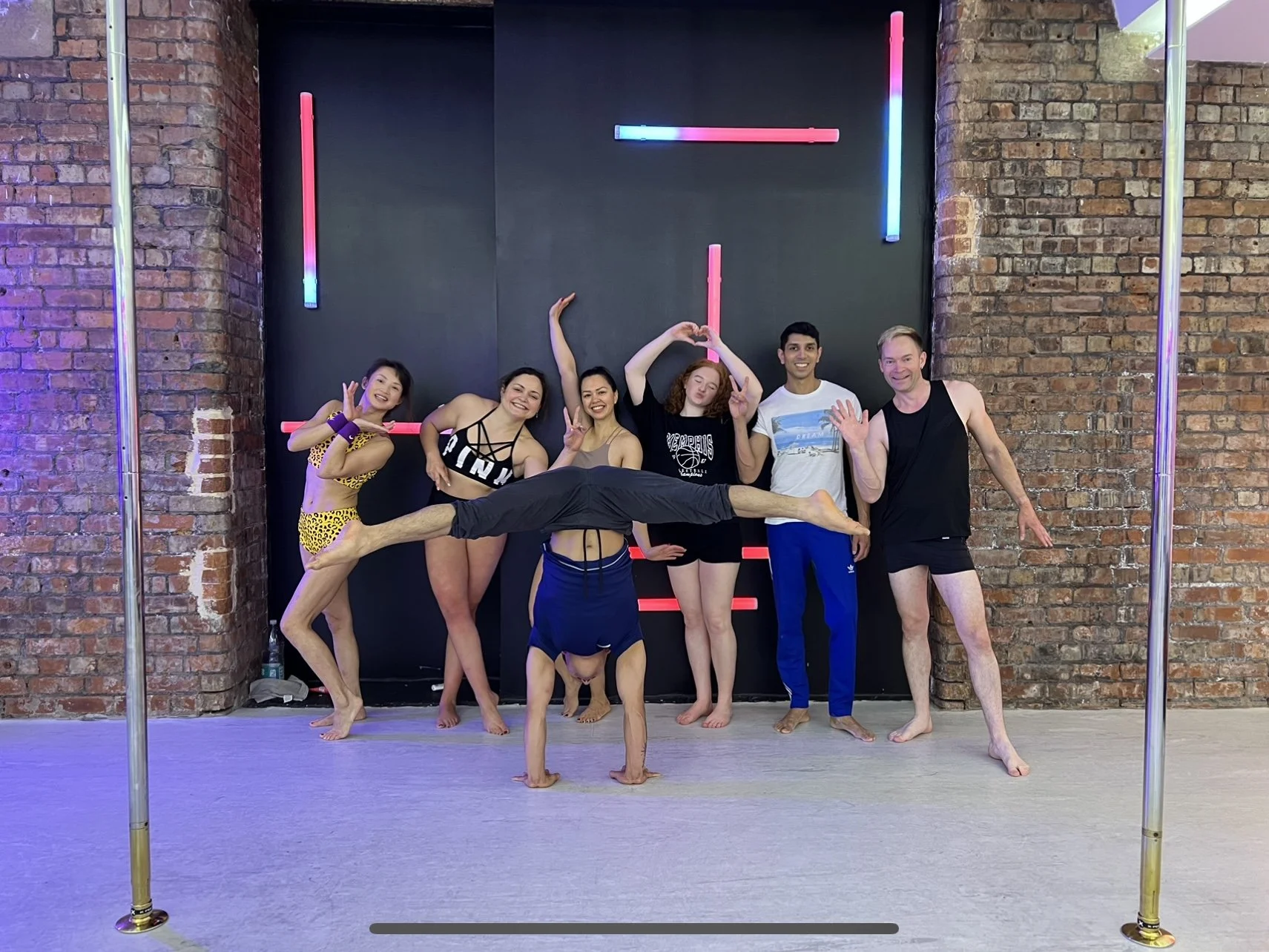 A group of seven people posing in a dance studio with brick walls and neon lights. One person is upside down in a handstand split, while the others stand in various poses. The scene appears lively and energetic.