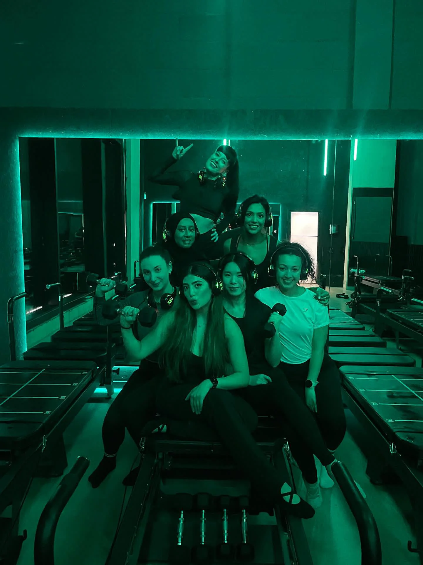Group of women with headphones in a gym, posing with weights, under green lighting.