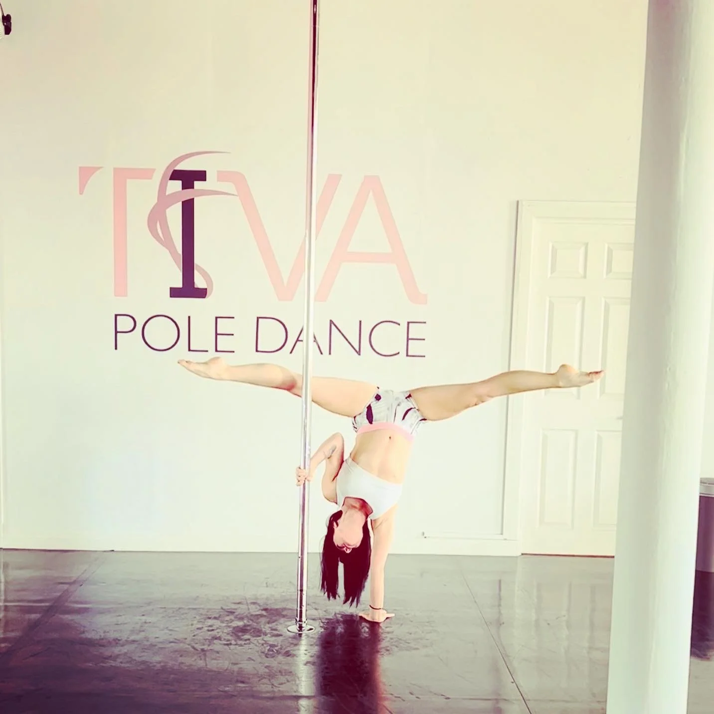 Person performing an advanced pole dance move in a studio with the words 'TIVA POLE DANCE' on the wall.